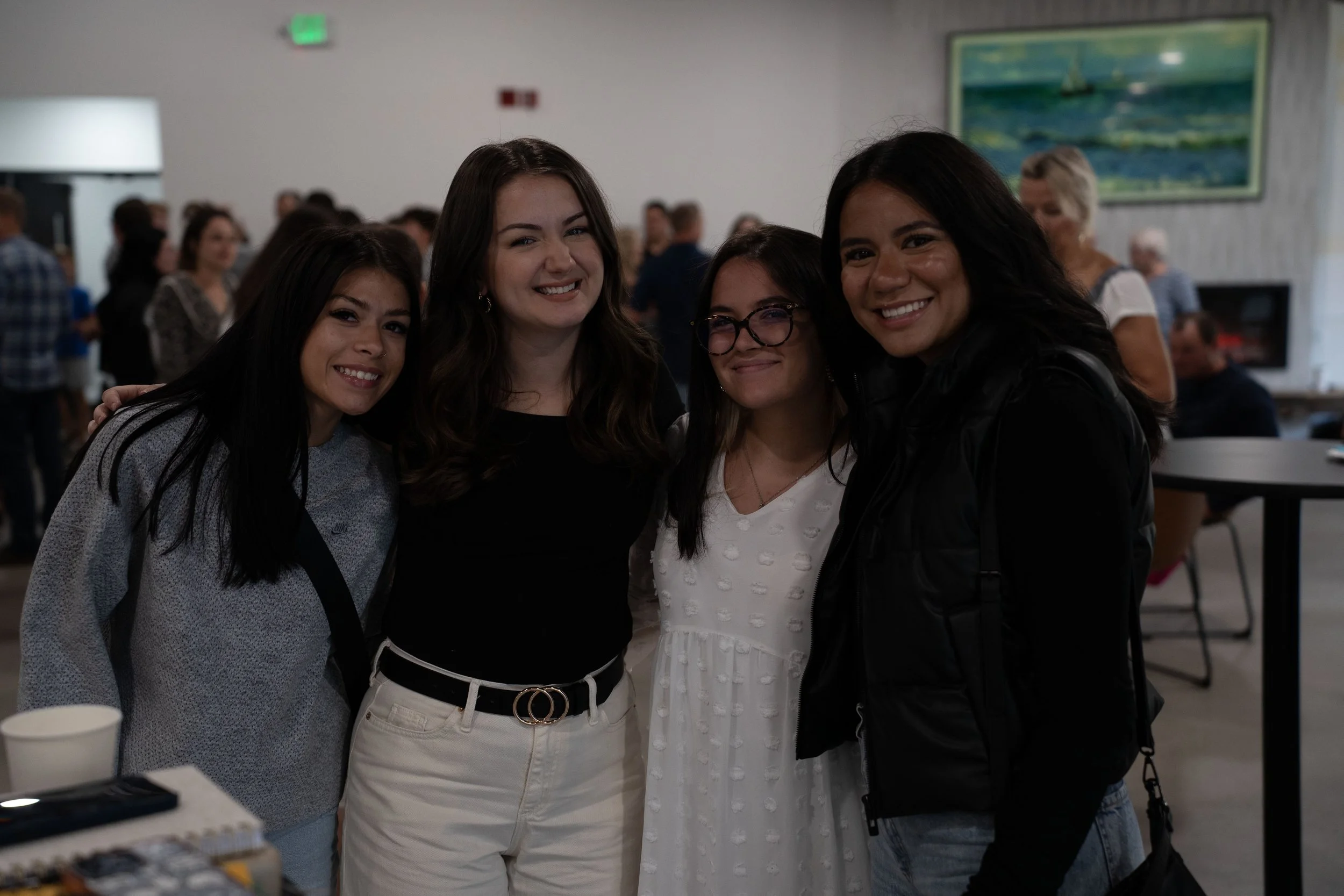 Four young women smiling together at an indoor social gathering, with people and artwork in the background.