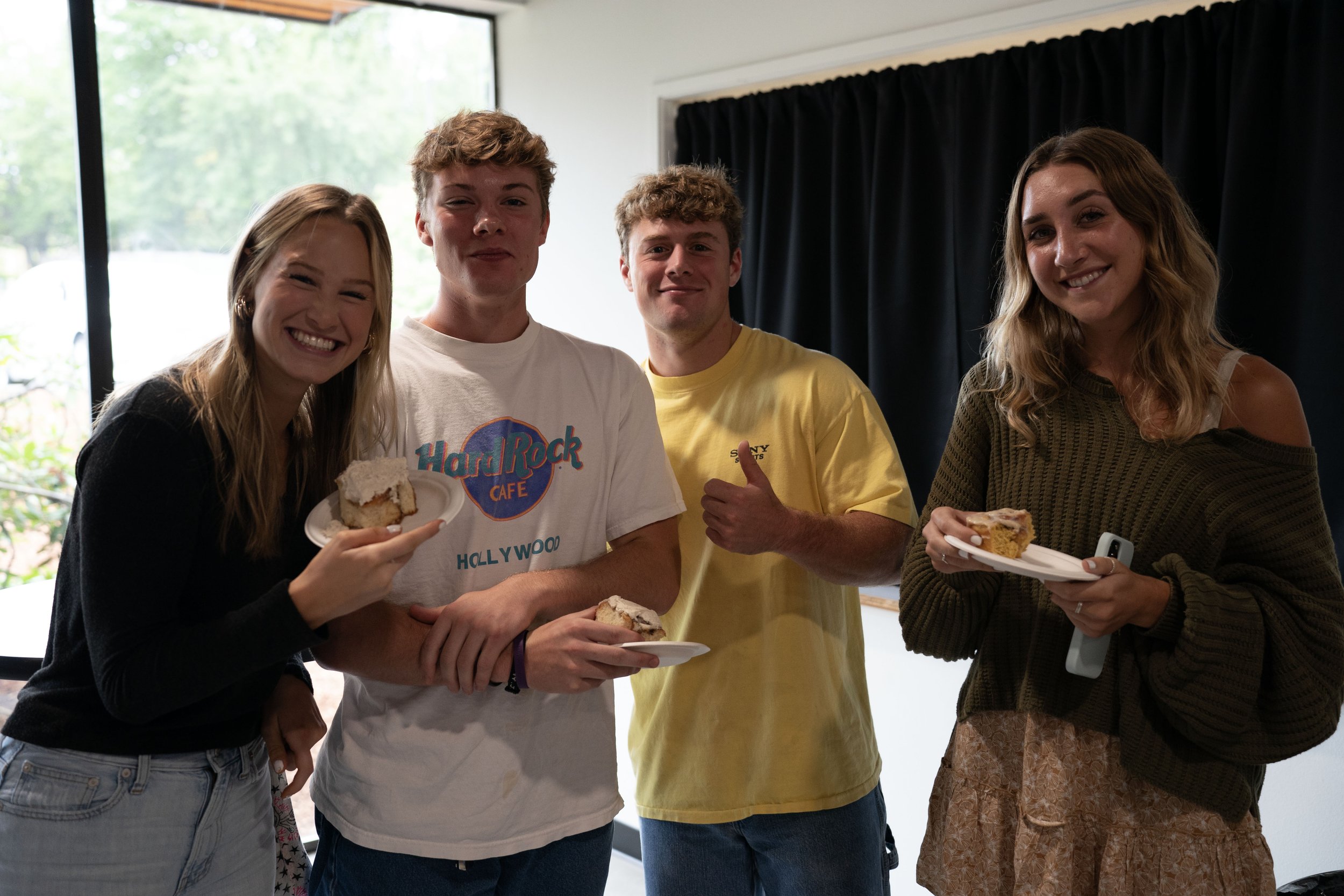 Four young adults standing indoors, two women and two men, holding plates with slices of cake and smiling at the camera.