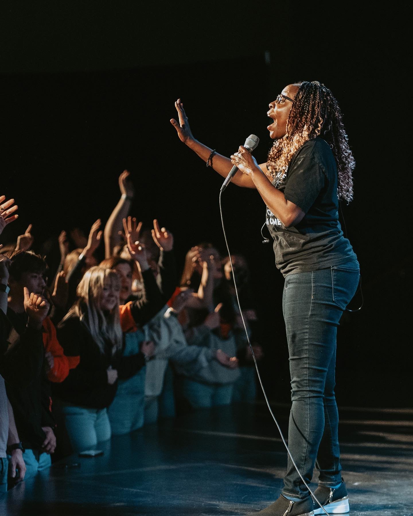 A woman speaking or singing on stage with a microphone, engaging with an audience of children who are raising their hands.