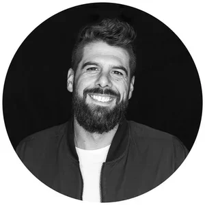 A smiling man with a beard and mustache, wearing a jacket, in a black and white circular portrait.