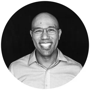 Black-and-white portrait of a smiling man with glasses, wearing a collared shirt, against a dark background.