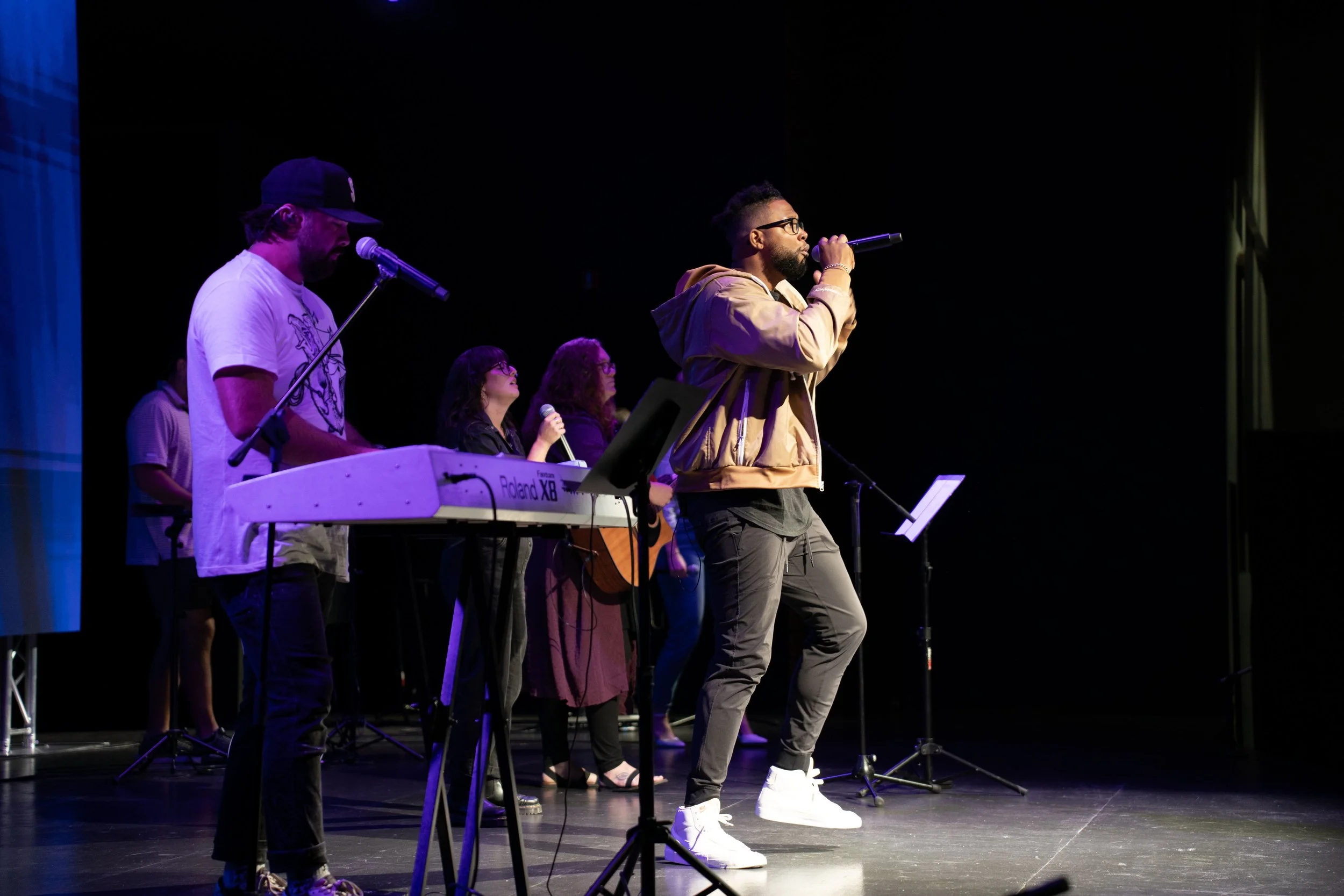 A group of five musicians performing on stage, including a singer with glasses and a beige jacket holding a microphone, a keyboard player, and three backup singers, with a dark background and stage lighting.