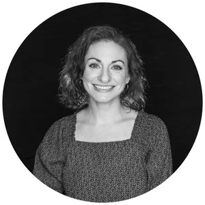 A black-and-white portrait of a smiling woman with curly hair, wearing a patterned top, against a dark circular background.