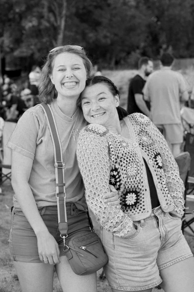 Two young women smiling and hugging outdoors at a gathering, with other people in the background.