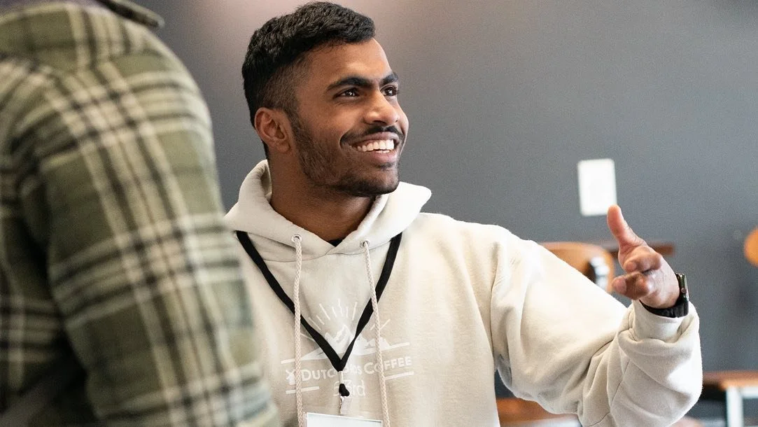 A smiling man with short dark hair and a beard wearing a beige hoodie with a logo, talking to another person in a plaid shirt in an indoor setting.
