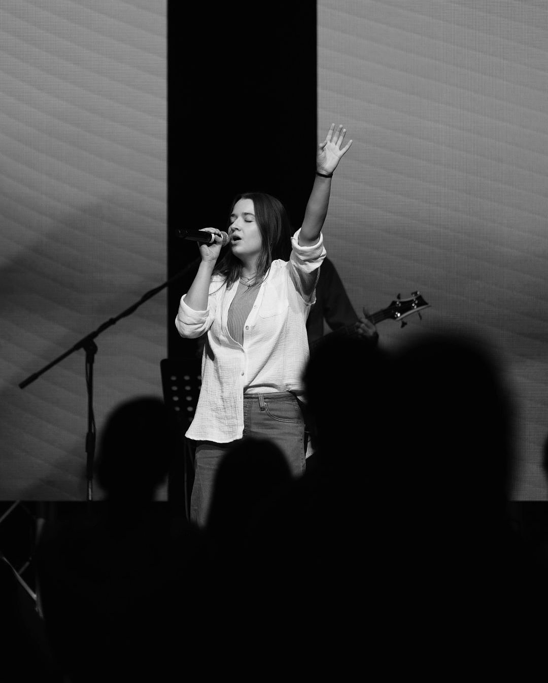 A woman singing into a microphone with her eyes closed, one arm raised, and a band playing behind her, in a live music performance.