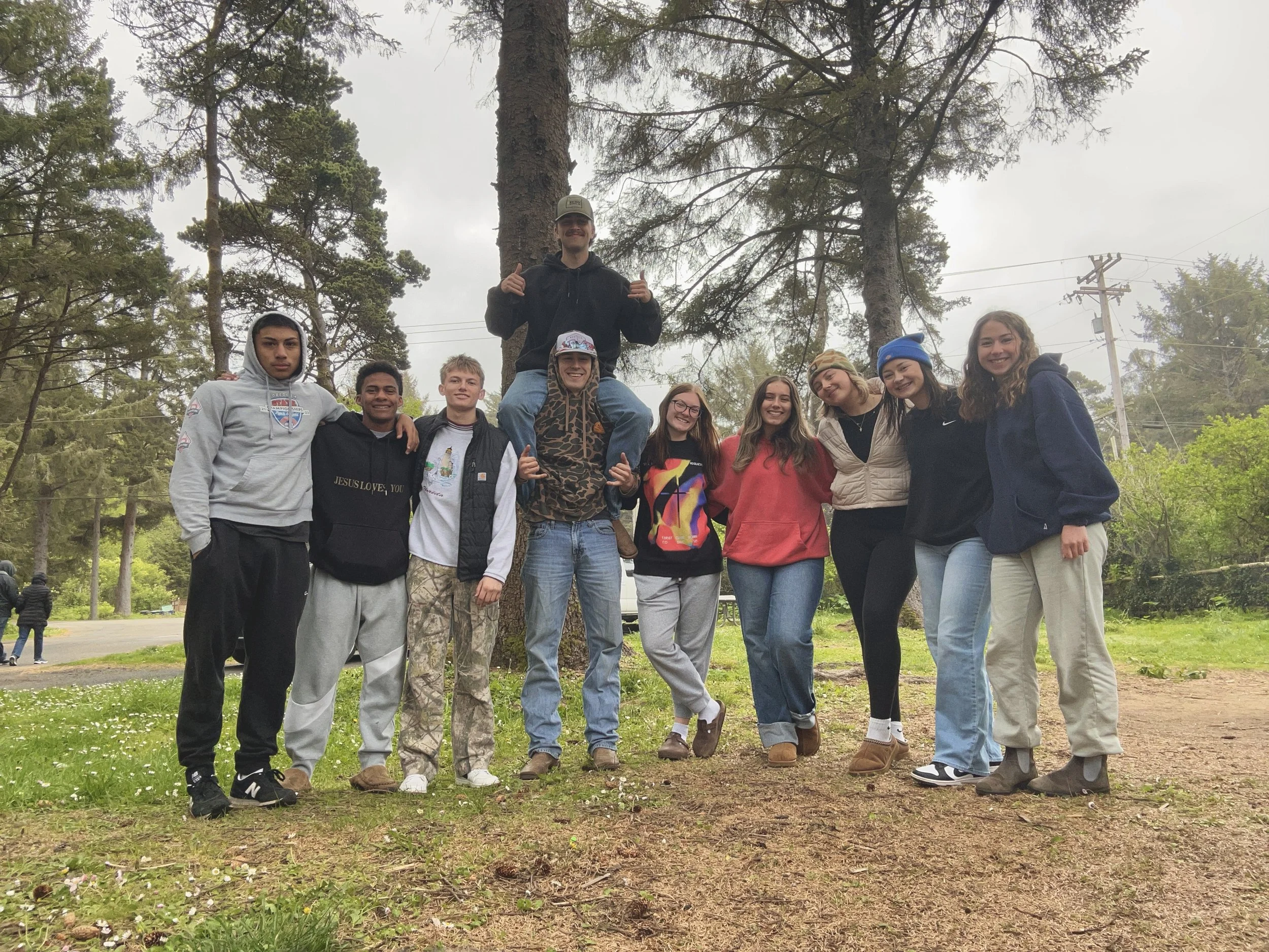 Group of nine young people standing together outdoors, some with their arms around each other, in front of trees on a cloudy day.