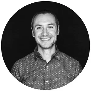Black and white portrait of a smiling man with short hair, wearing a dotted shirt, against a dark background.