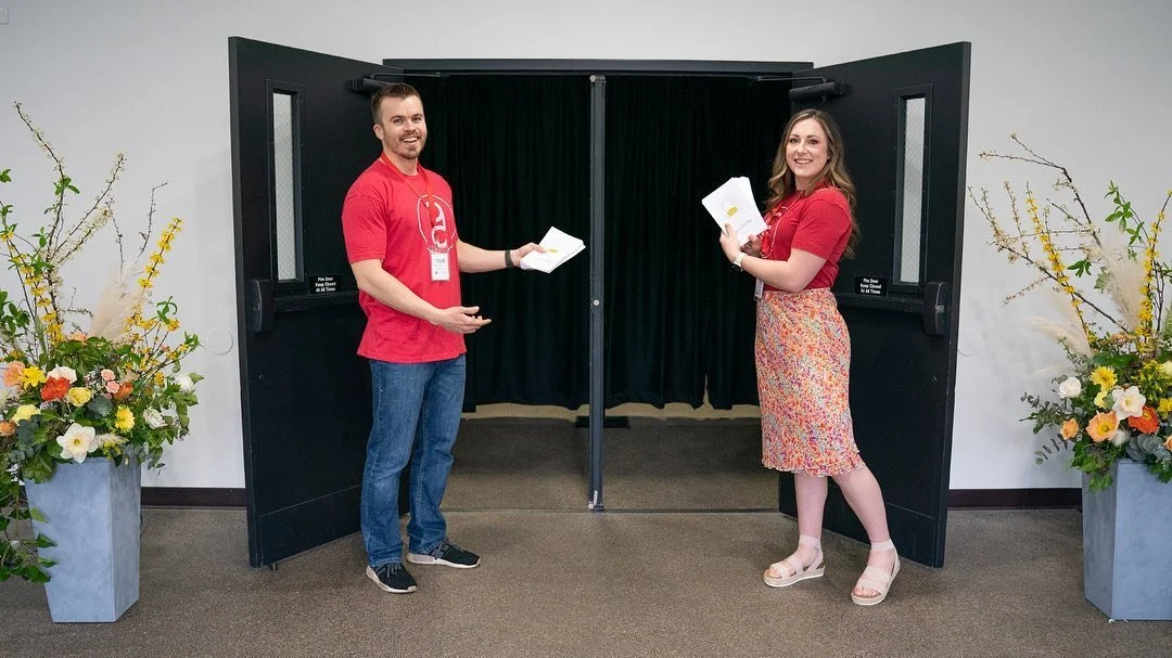 Two smiling people holding papers in front of a black photo booth with closed curtains, flanked by two large flower arrangements in gray pots.