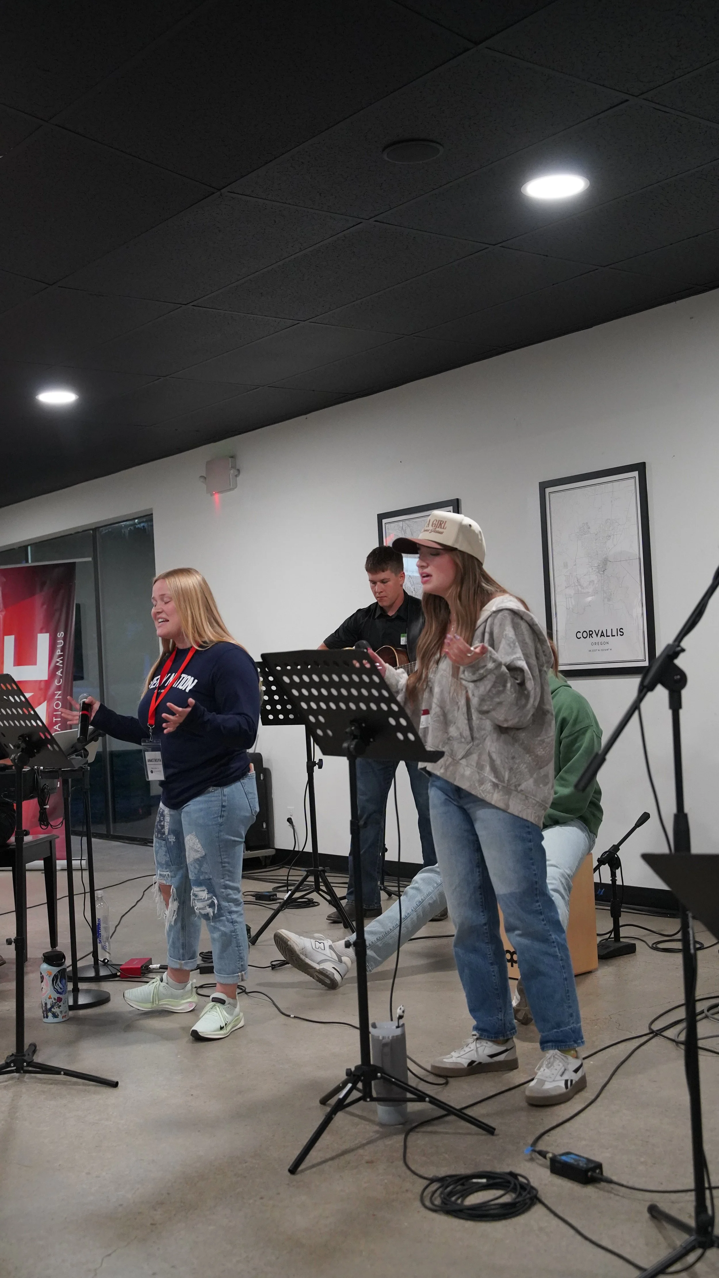 Group of young people singing and playing instruments during a performance indoors, with music stands, microphones, and a guitar.