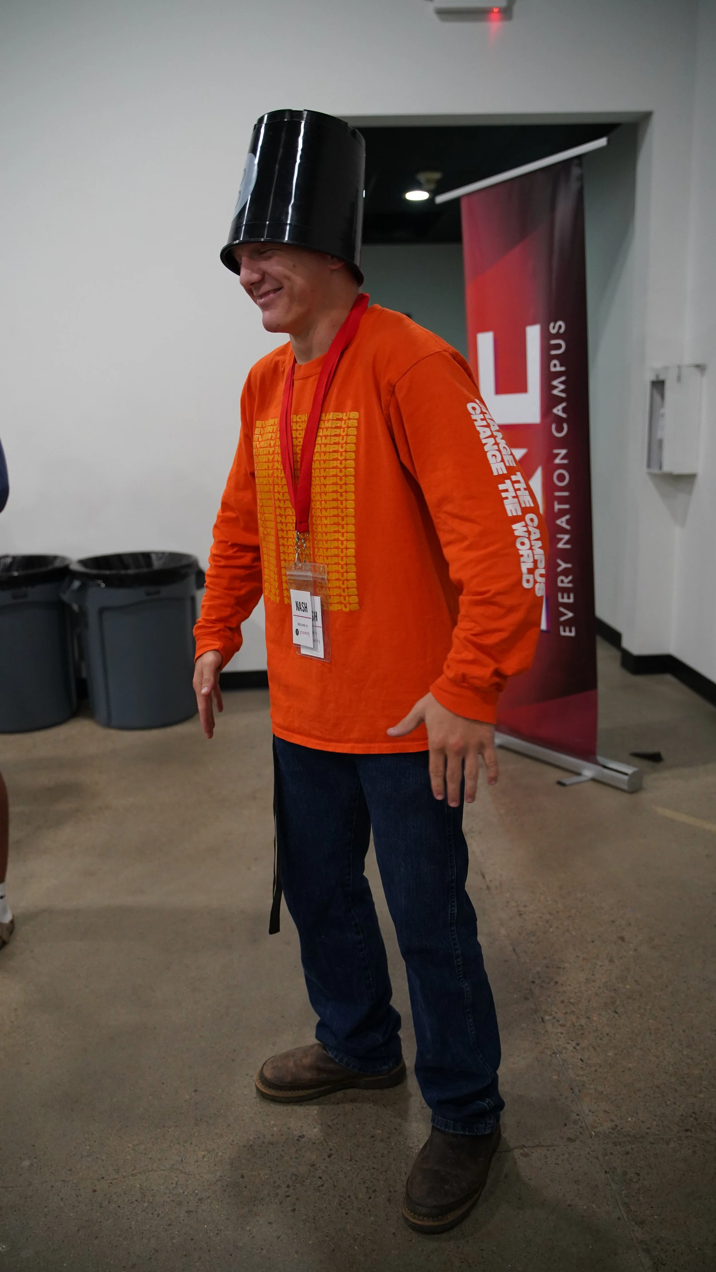 A man wearing a black trash can on his head, an orange long-sleeve shirt, and jeans, standing with a cheerful expression inside a room with a banner in the background.