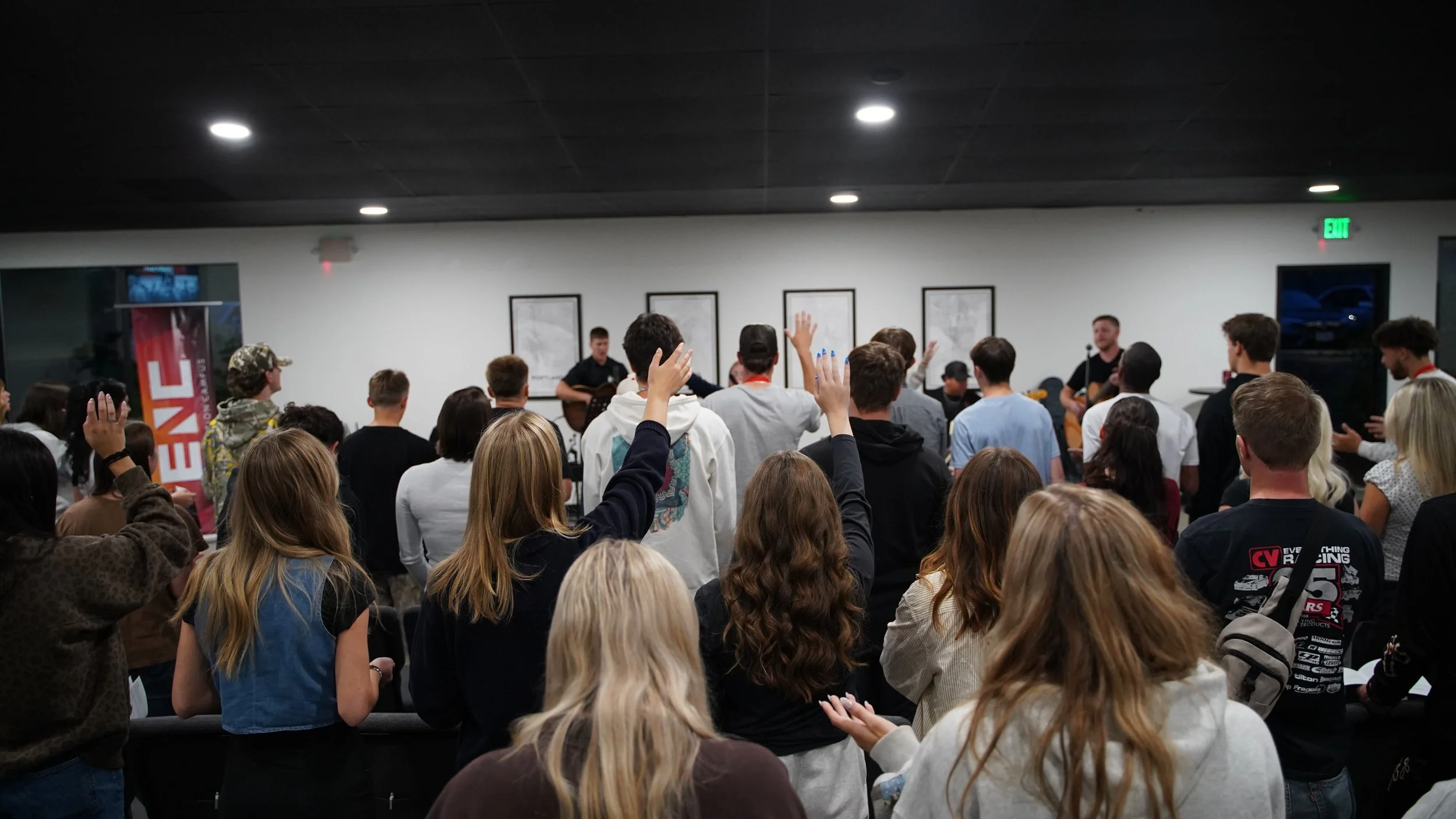 Group of people gathered indoors, some raising hands, listening to musicians playing guitars on stage with framed artwork on the wall behind.