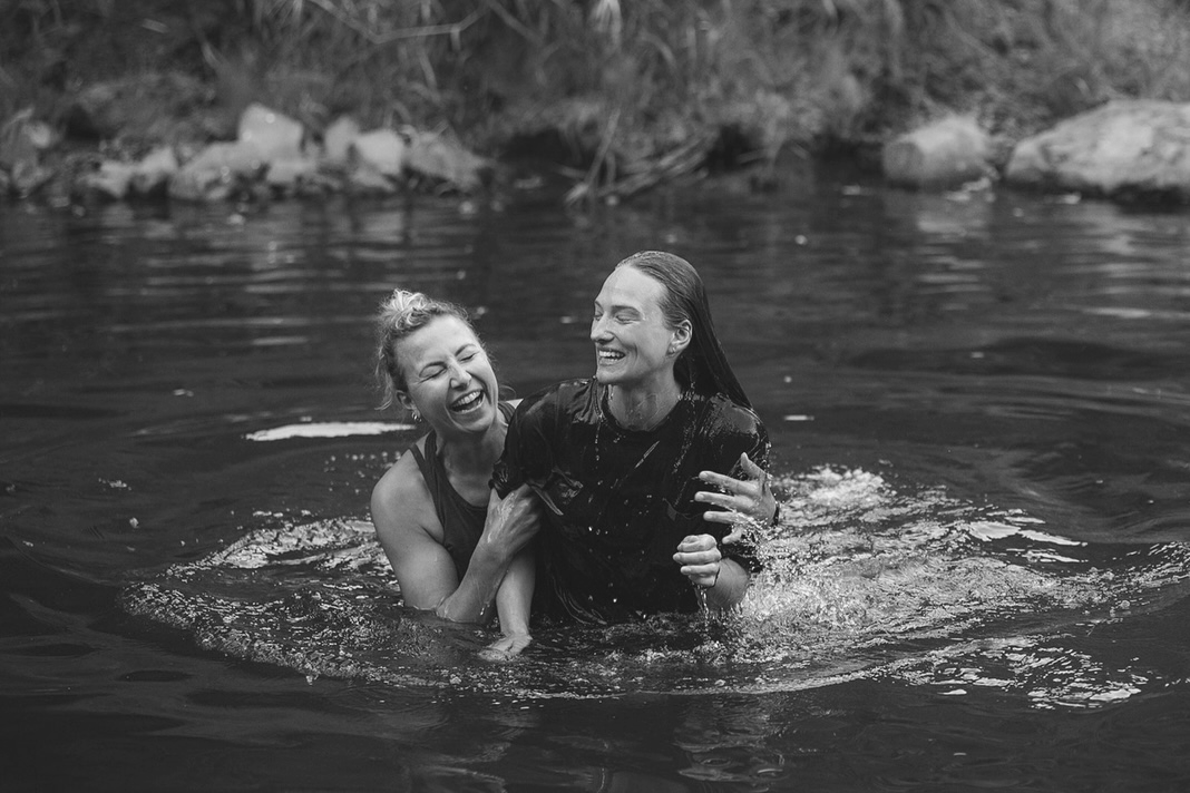 Two women laughing and playing in a body of water, with one woman helping the other to stand, surrounded by rocks and grass.