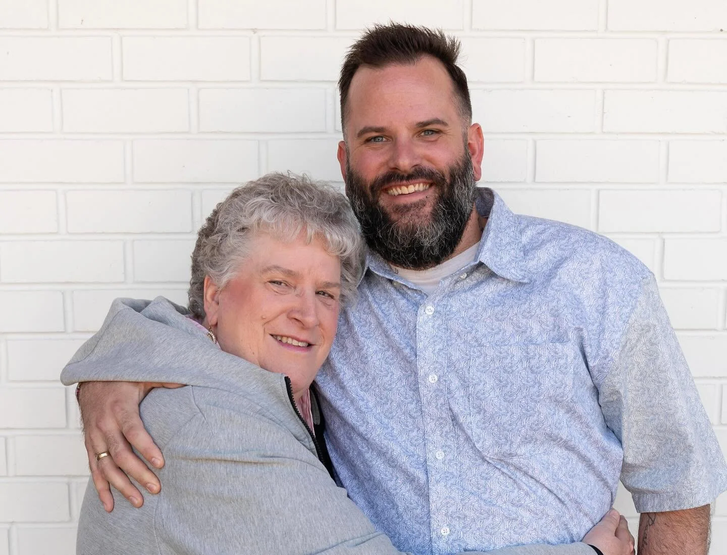 A smiling elderly woman embracing a man with a beard, both standing against a white brick wall, in a friendly and affectionate pose.