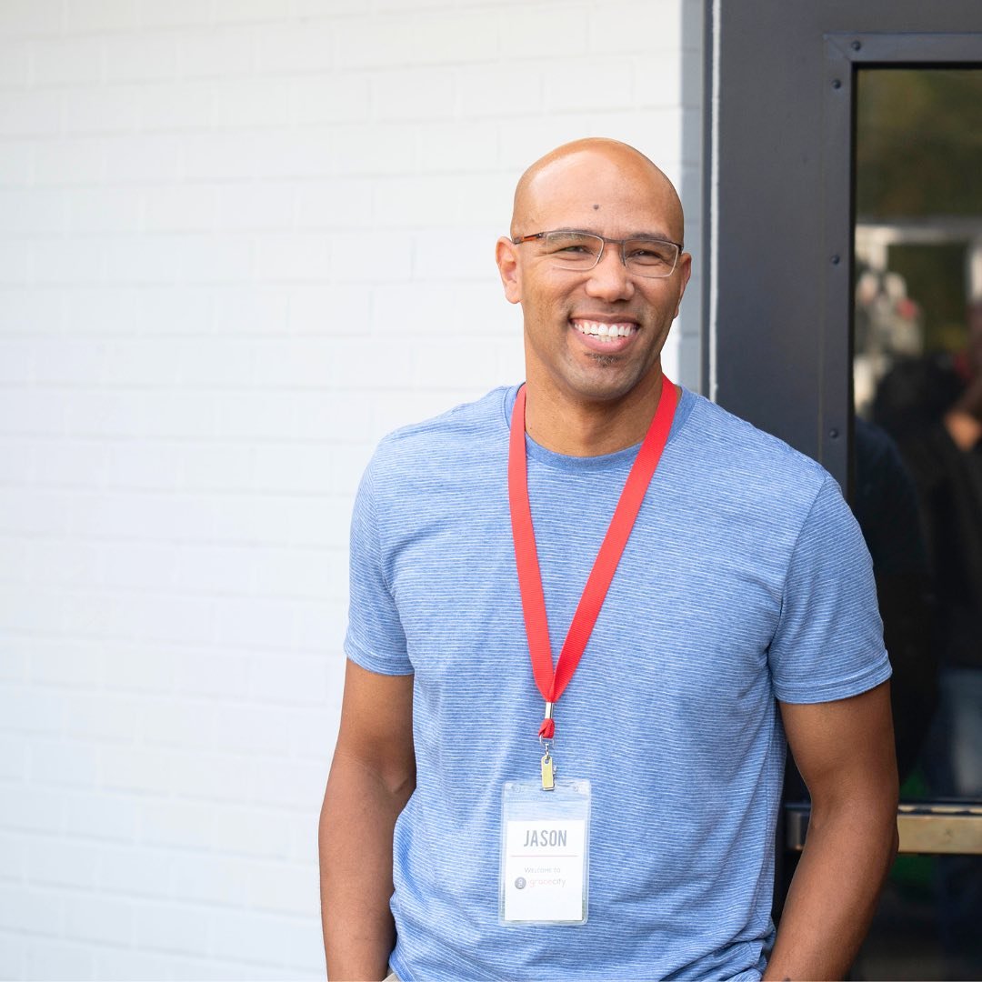 A smiling man with glasses and a bald head, wearing a blue t-shirt and a red lanyard with a name tag that reads 'Jason,' standing outside near a white brick wall and a black door.