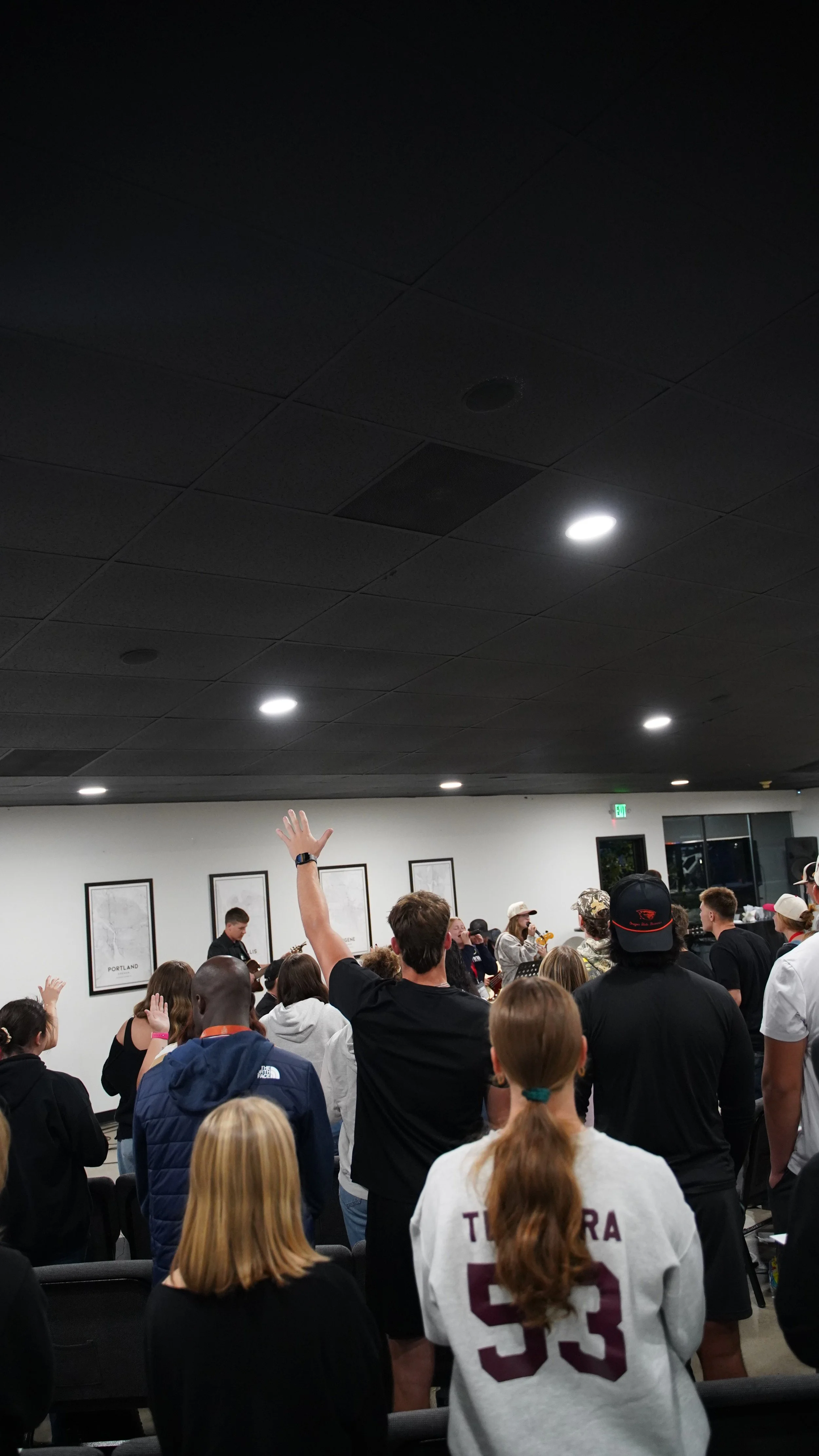 People gathered indoors, some raising their hands, listening to someone speaking or performing in front of framed maps or posters on the wall.