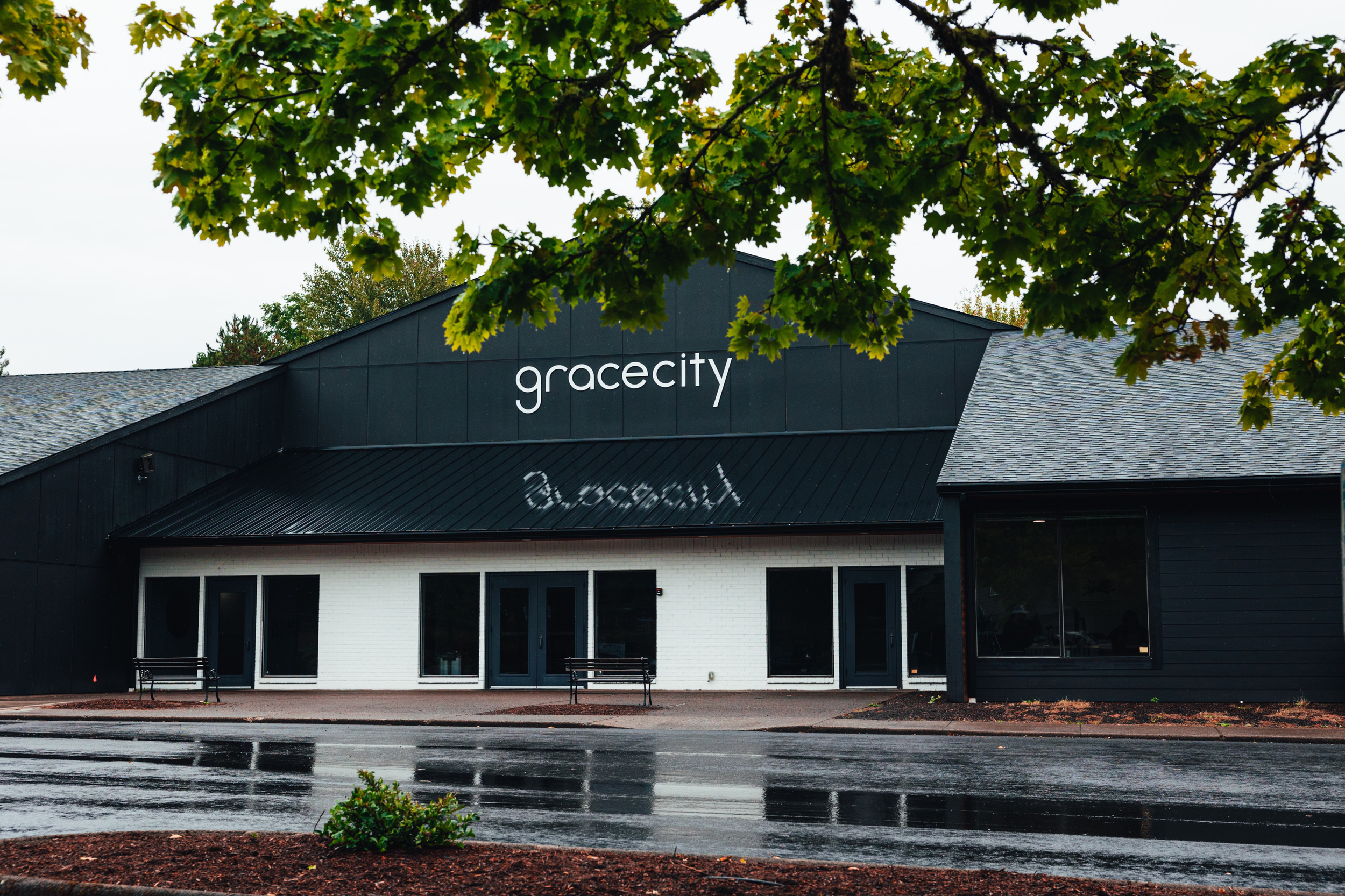 The Grace City building with black and white exterior, black roof, and the name 'gracecity' displayed on the front, wet pavement and trees in the foreground and overcast skies.