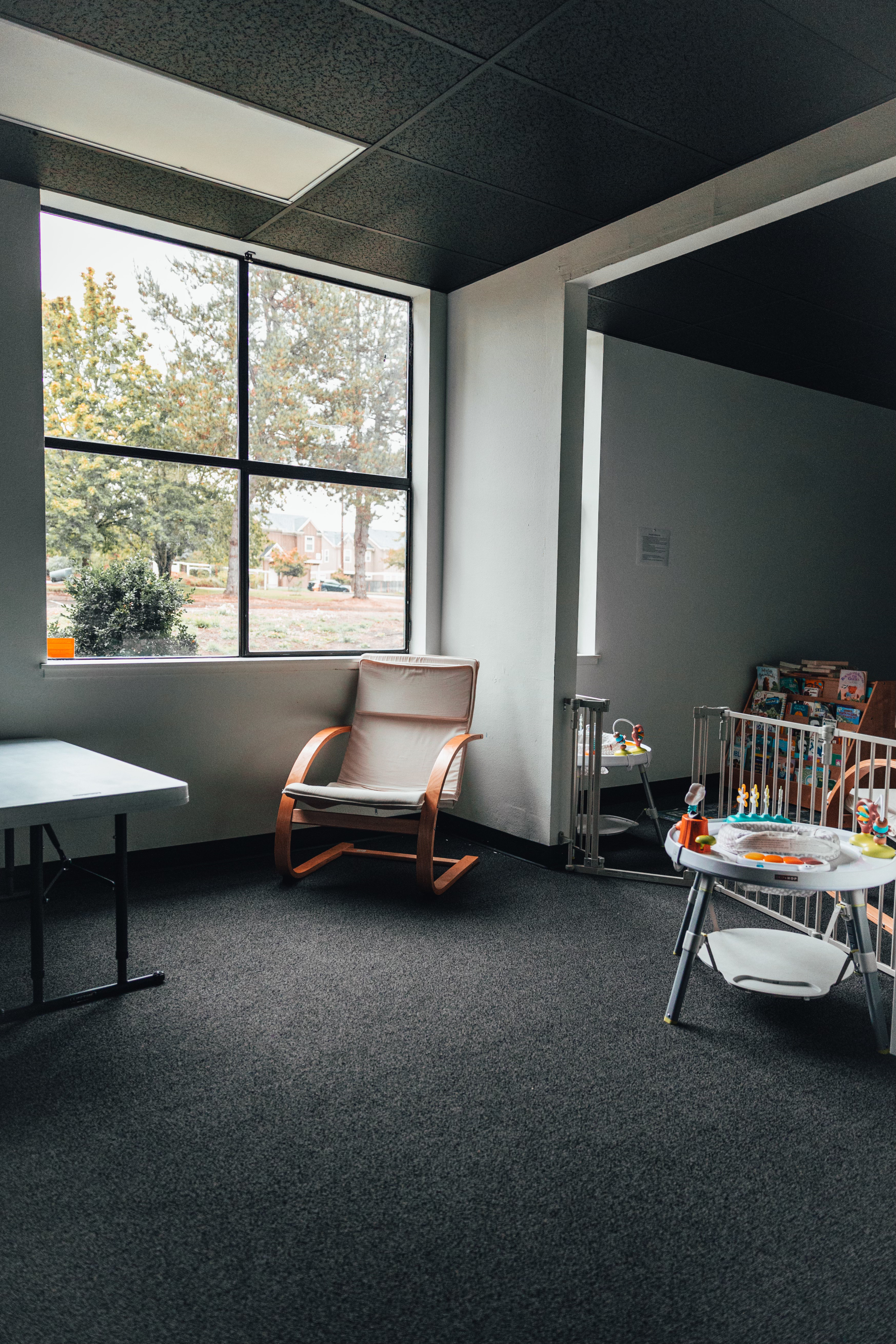 An empty room with a large window, a beige armchair by the window, a table, and a baby play area with toys and books.