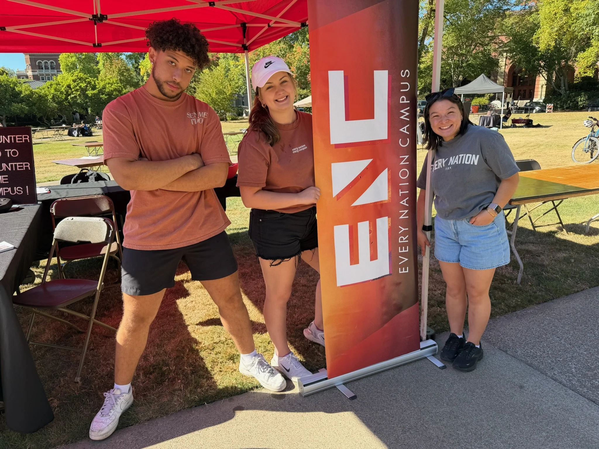 Three young adults standing outdoors near a red canopy with a sign that reads "EVEL U" at an Everett Community College event. Two women and one man are smiling and posing for the photo, with tables and other tents in the background on a sunny day.
