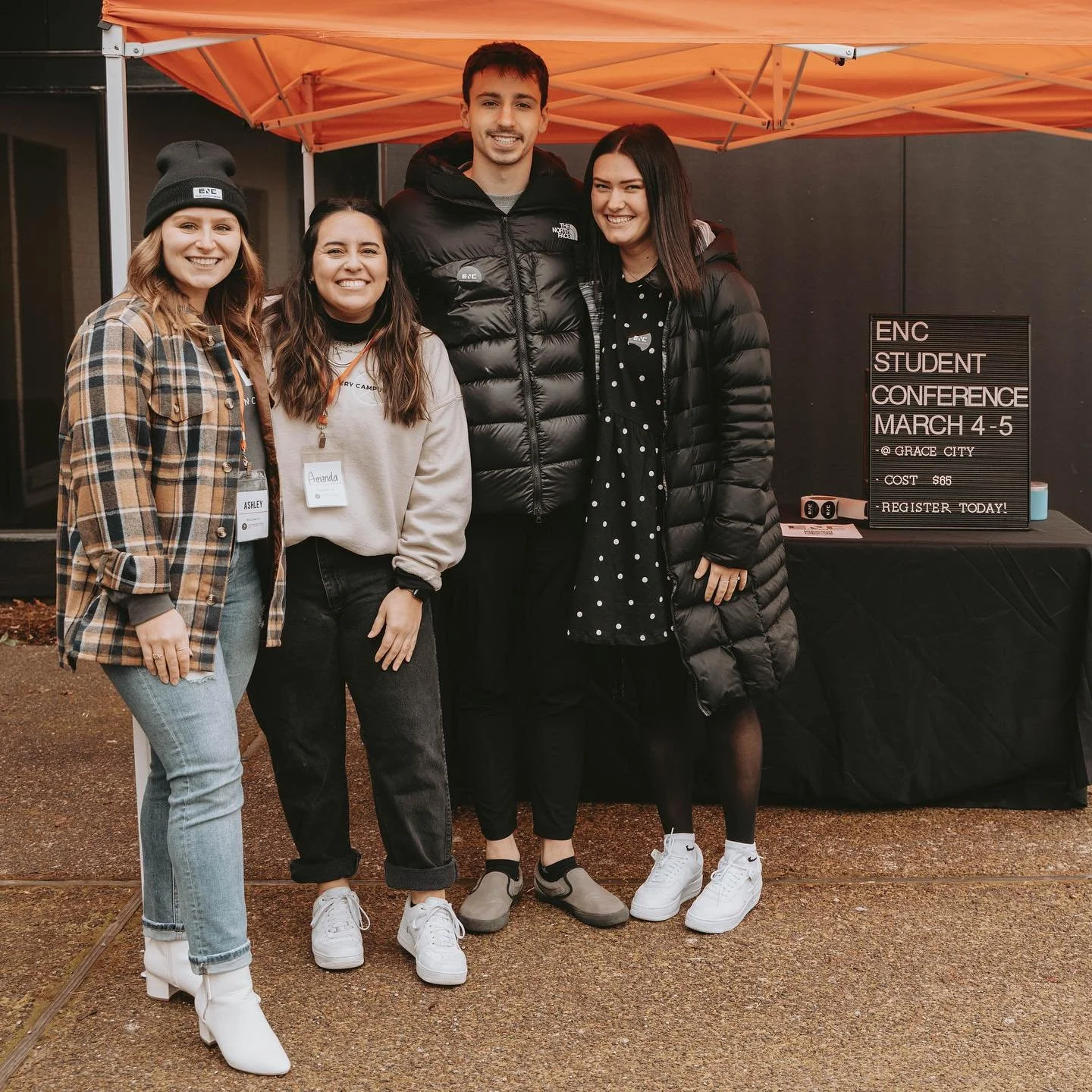 Four young women standing under an orange canopy at an outdoor event, smiling for the camera. There is a table with a sign promoting an upcoming student conference in the background.