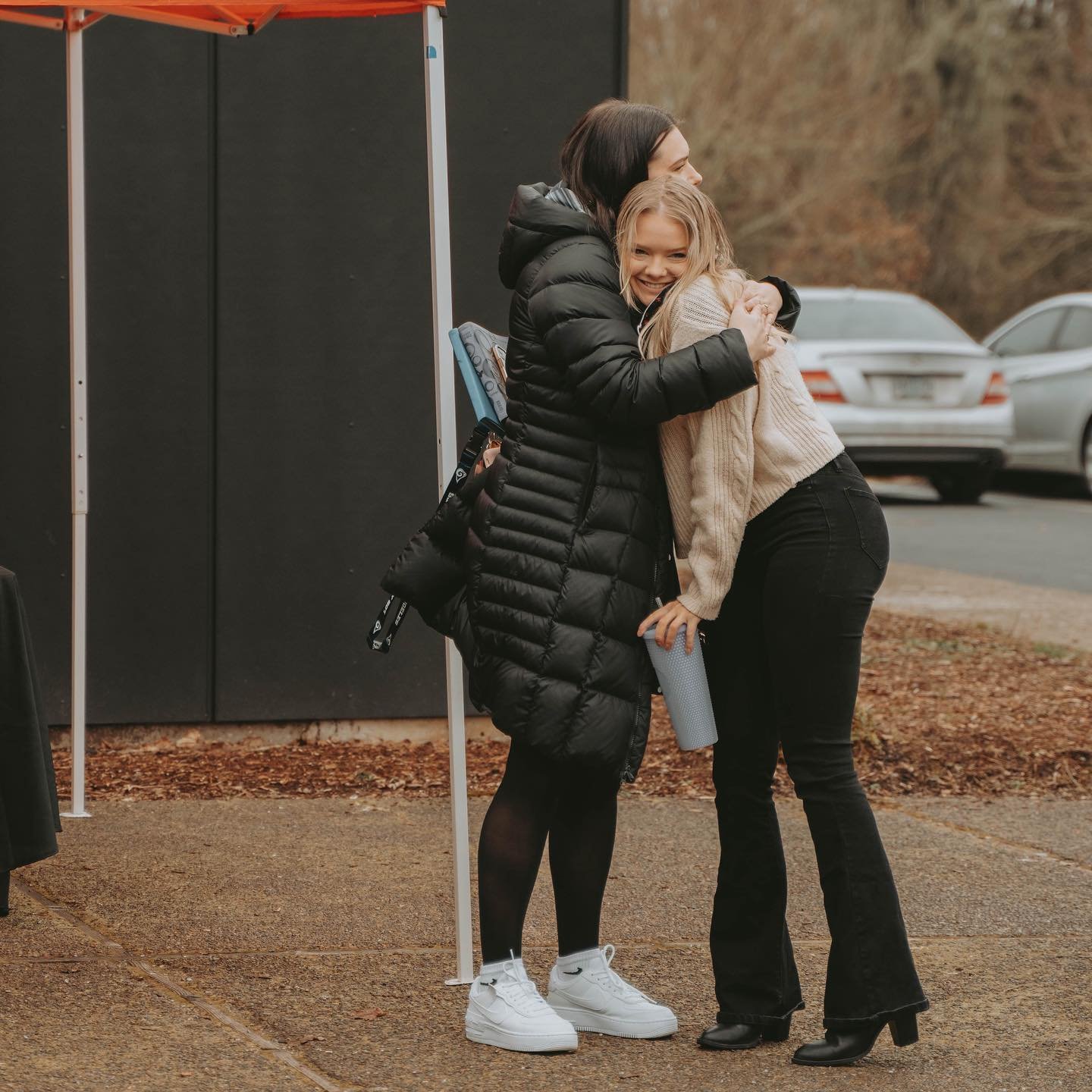 Two women hugging outdoors on a cloudy day near parked cars and an orange tent.