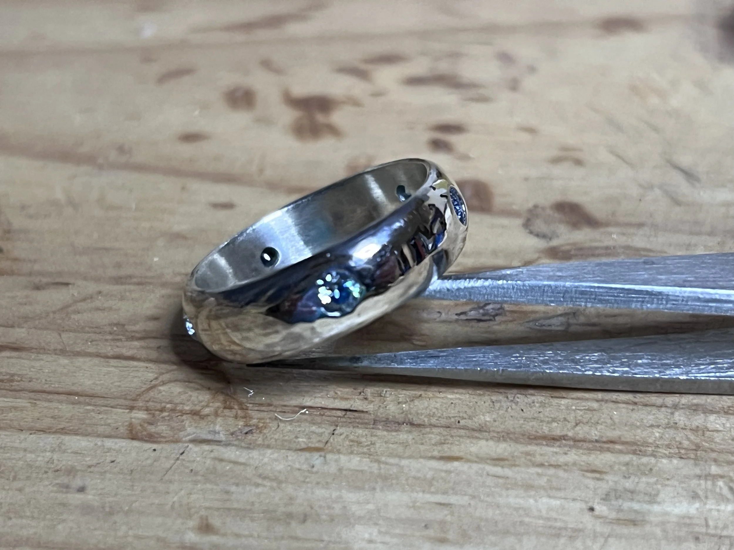 Close-up of a silver ring with embedded small blue gemstones, resting on a wooden surface, held by a metallic tool.