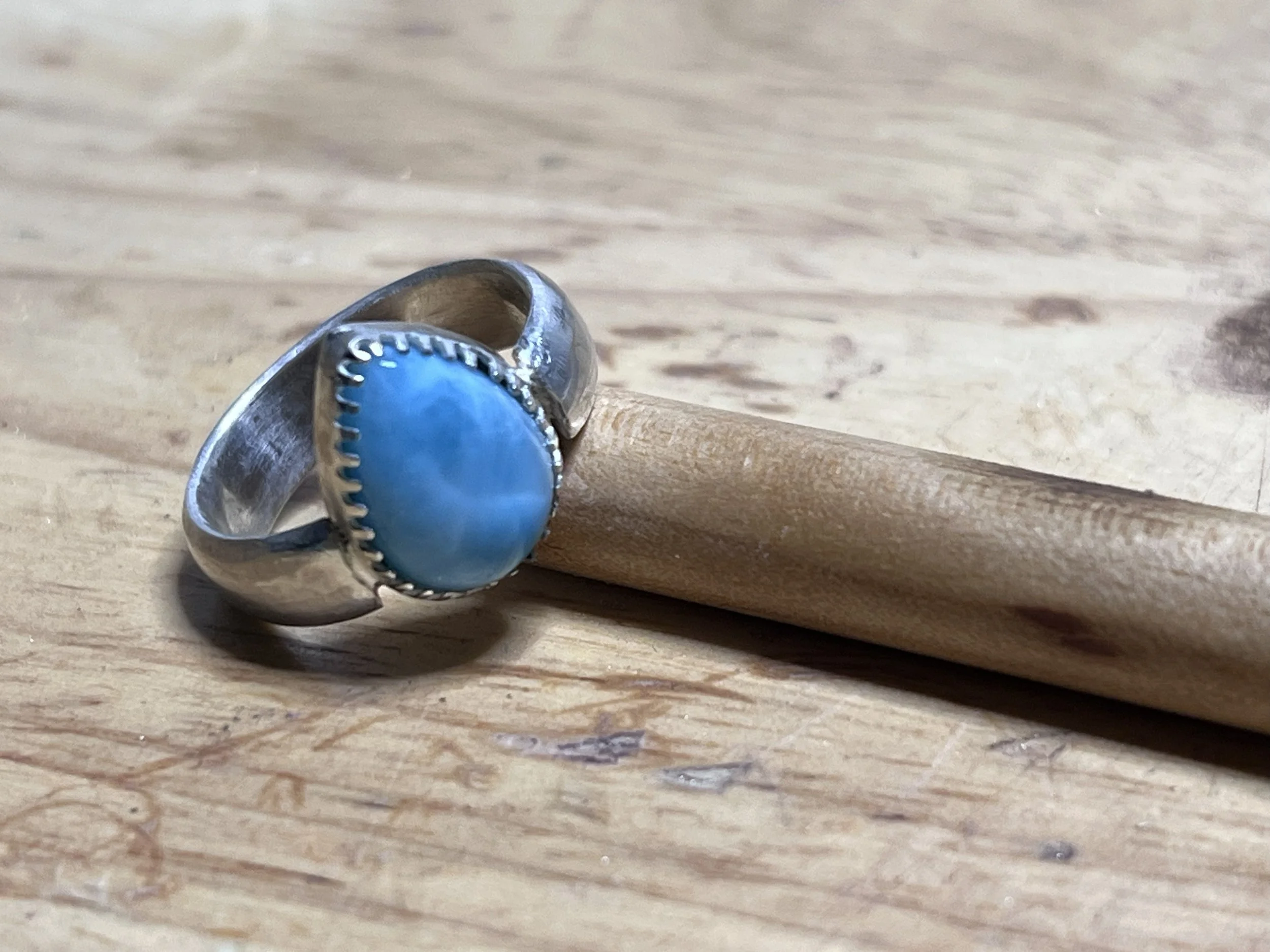 A silver ring with a large oval turquoise stone, resting on a wooden surface with a small wooden stick underneath.