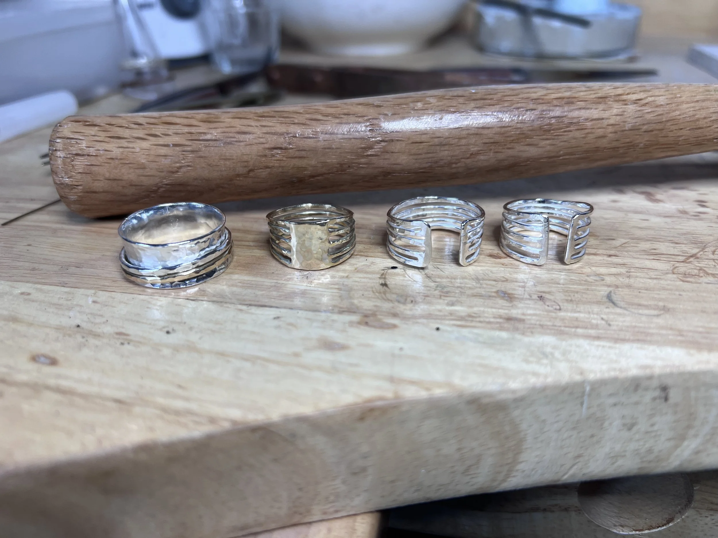 Three silver rings, 2 open style rings, one gold ring on a wooden work surface, with a wooden mallet in the background.