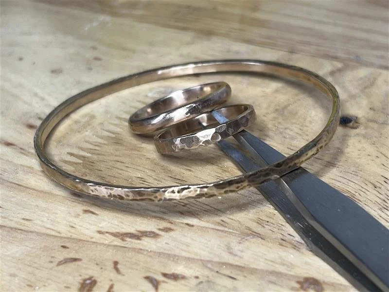A silver bracelet, two silver rings, and a flat metal tool resting on a wooden surface.