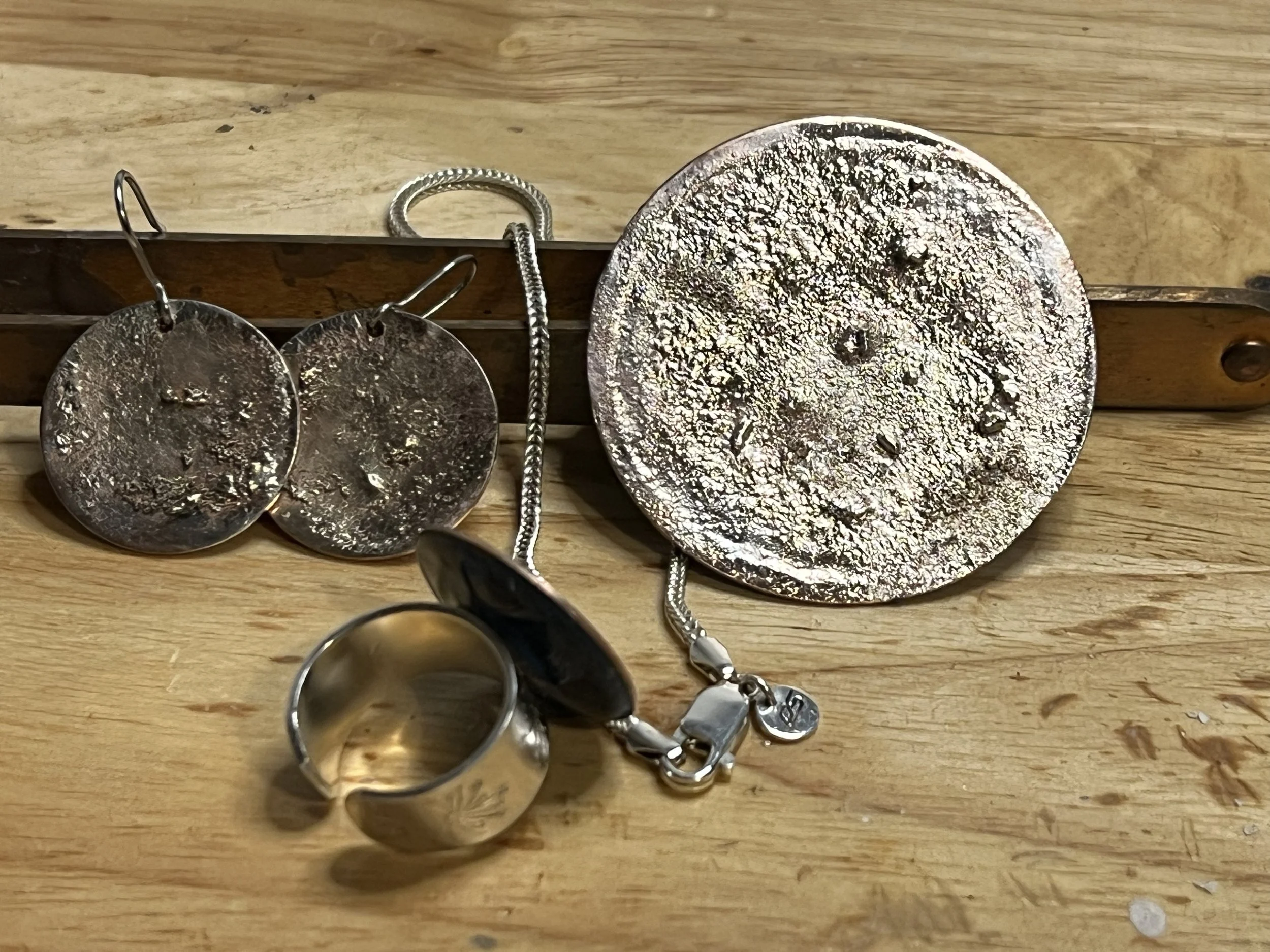 Silver jewelry including a large circular pendant, a pair of circular earrings, a chunky ring, and a necklace, displayed on a wooden surface with a wooden backing.