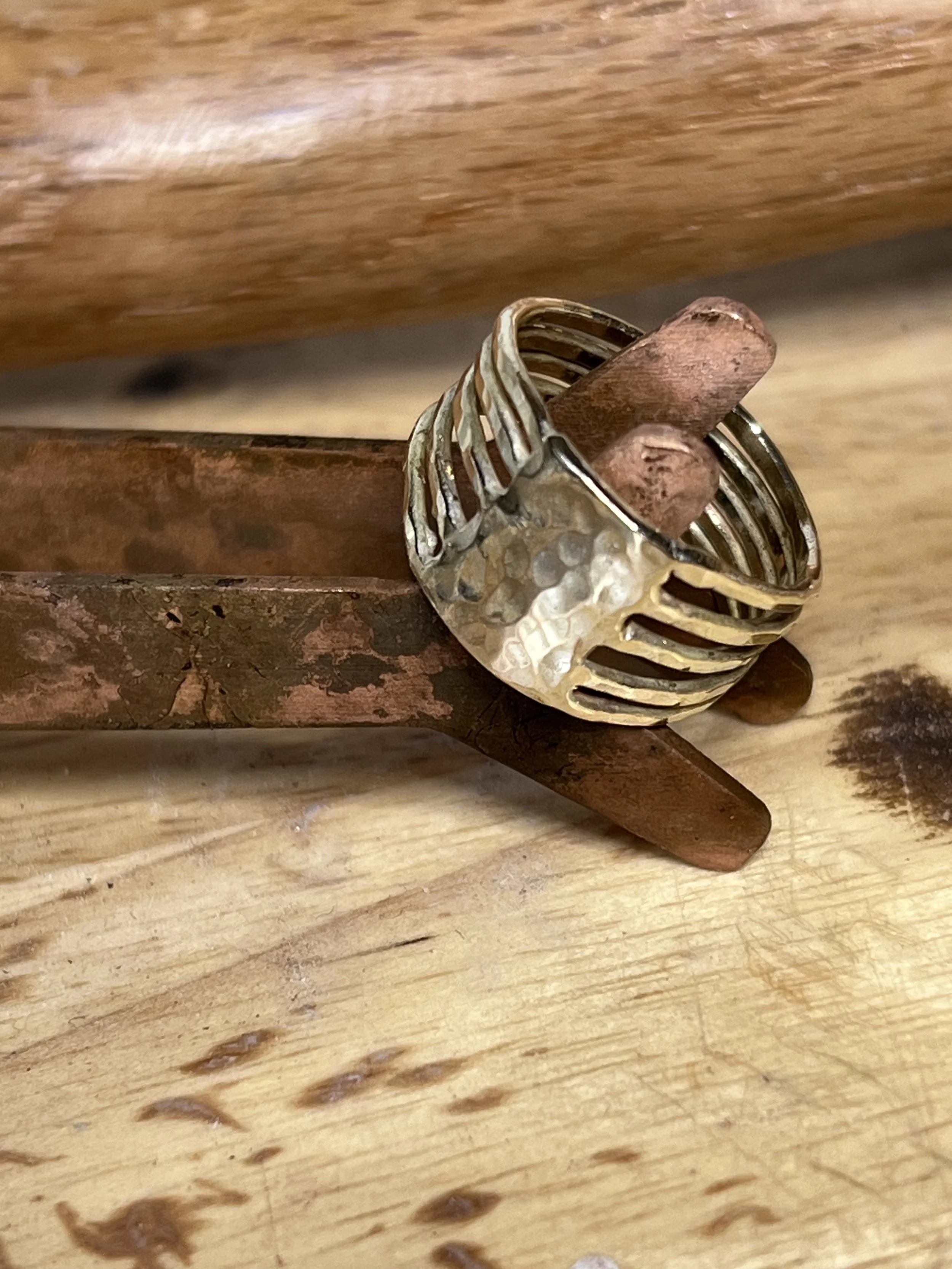 A gold ring with an open, lattice design, resting on a pair of copper tongs, on a wooden surface.