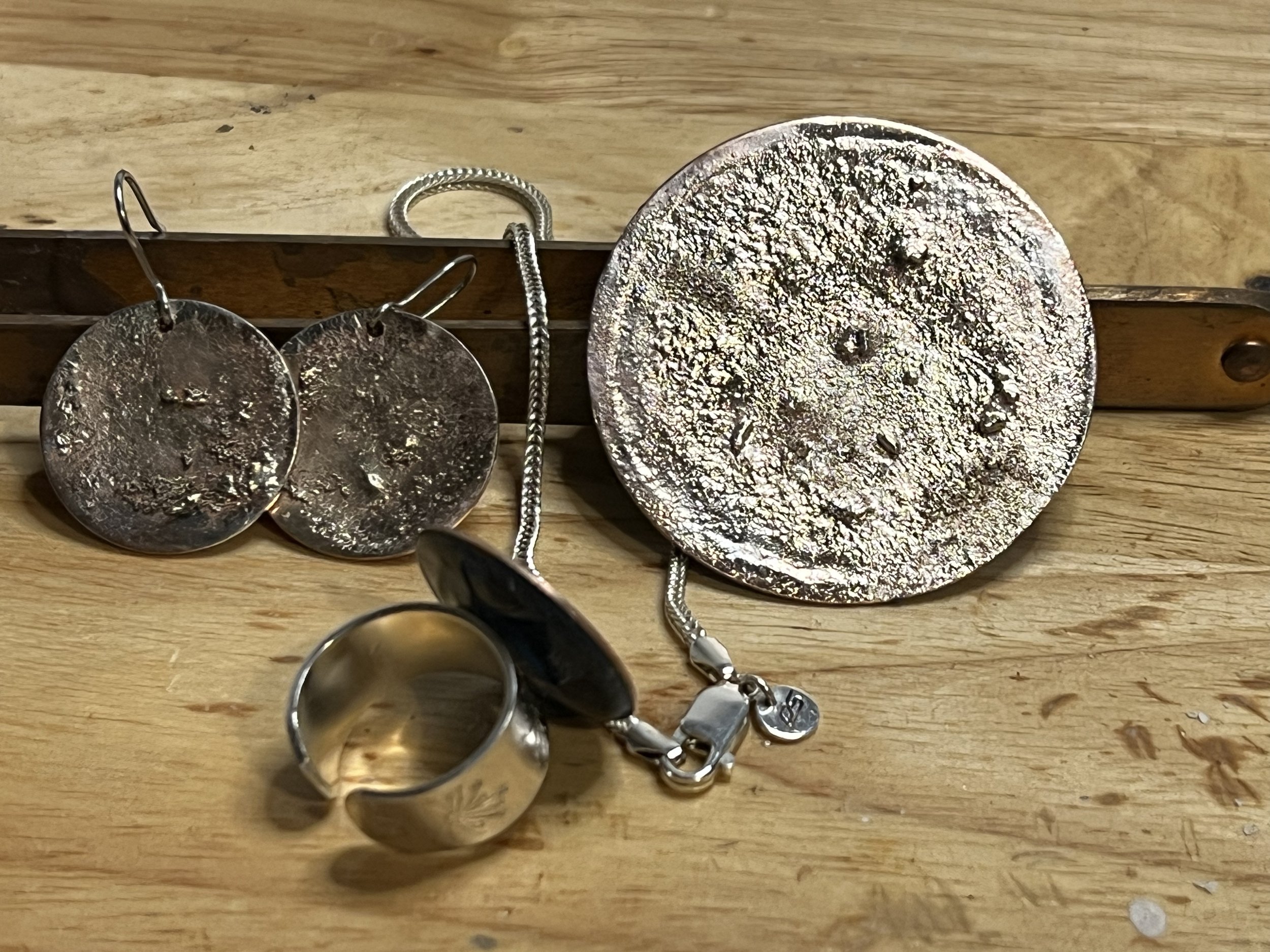 Silver jewelry including a wide ring, a pair of round earrings, large circular pendant, and a chain, arranged on a wooden surface.