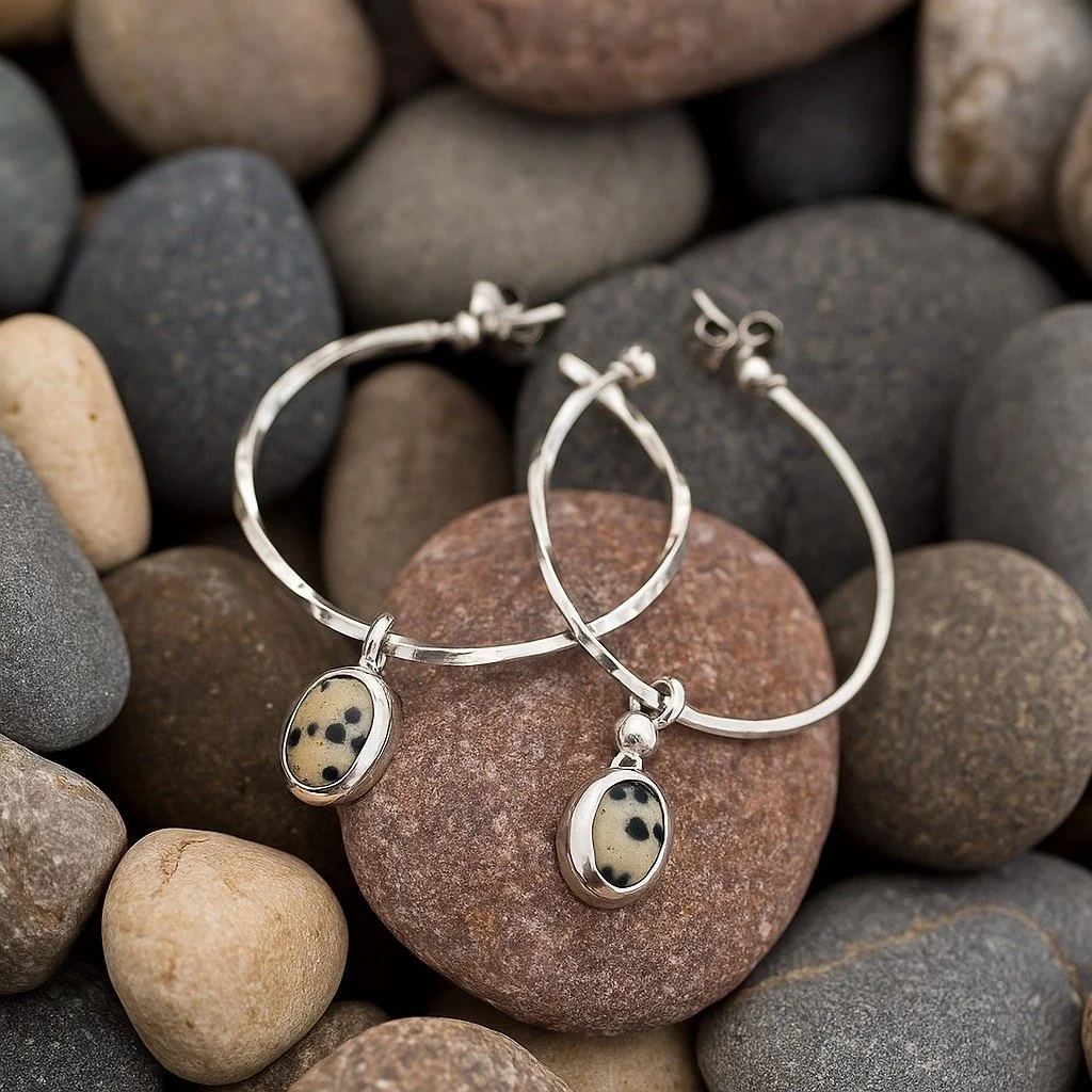 Silver hoop earrings with oval dalmatian jasper stones resting on multicolored smooth pebbles.