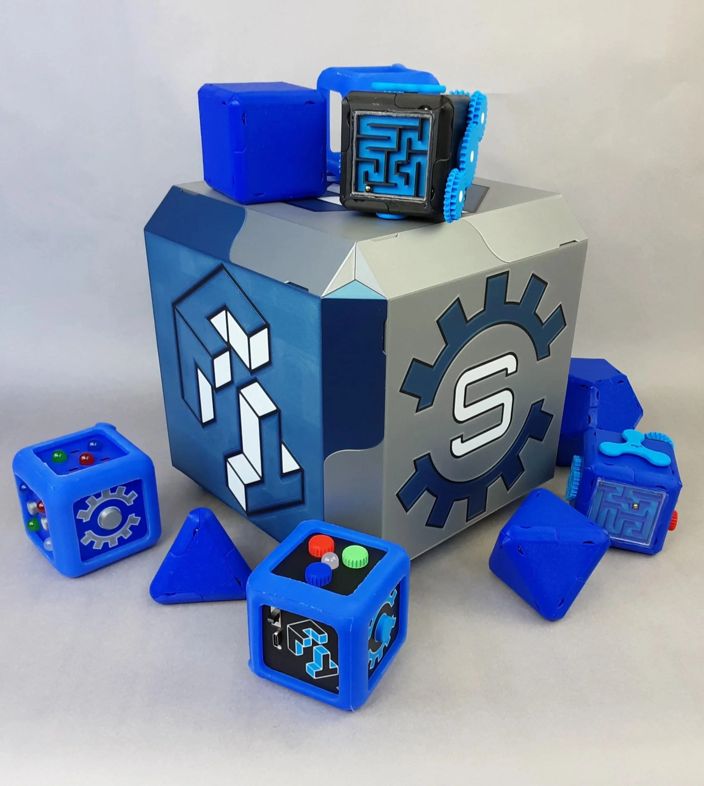 A robot-themed toy set featuring a large gray box with gear and gear-socket designs, surrounded by smaller blue cubes with gear patterns and control panels, all in shades of blue, gray, and black.
