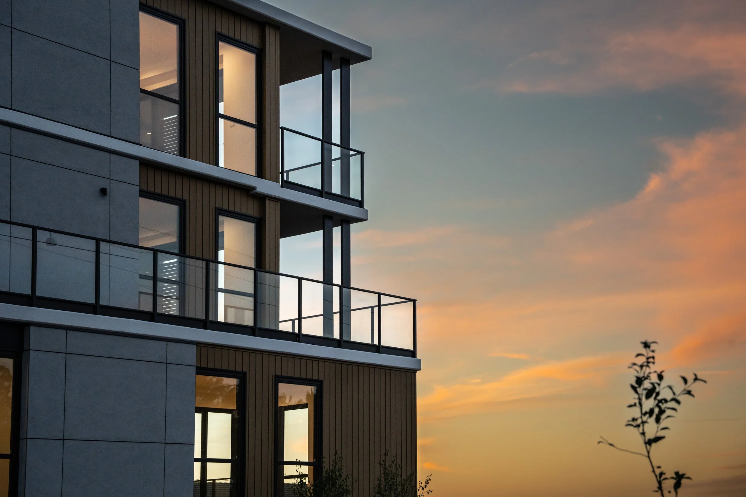 Modern multi-story apartment building with glass balconies and large windows at sunset, with a small tree in the foreground and a colorful sky.