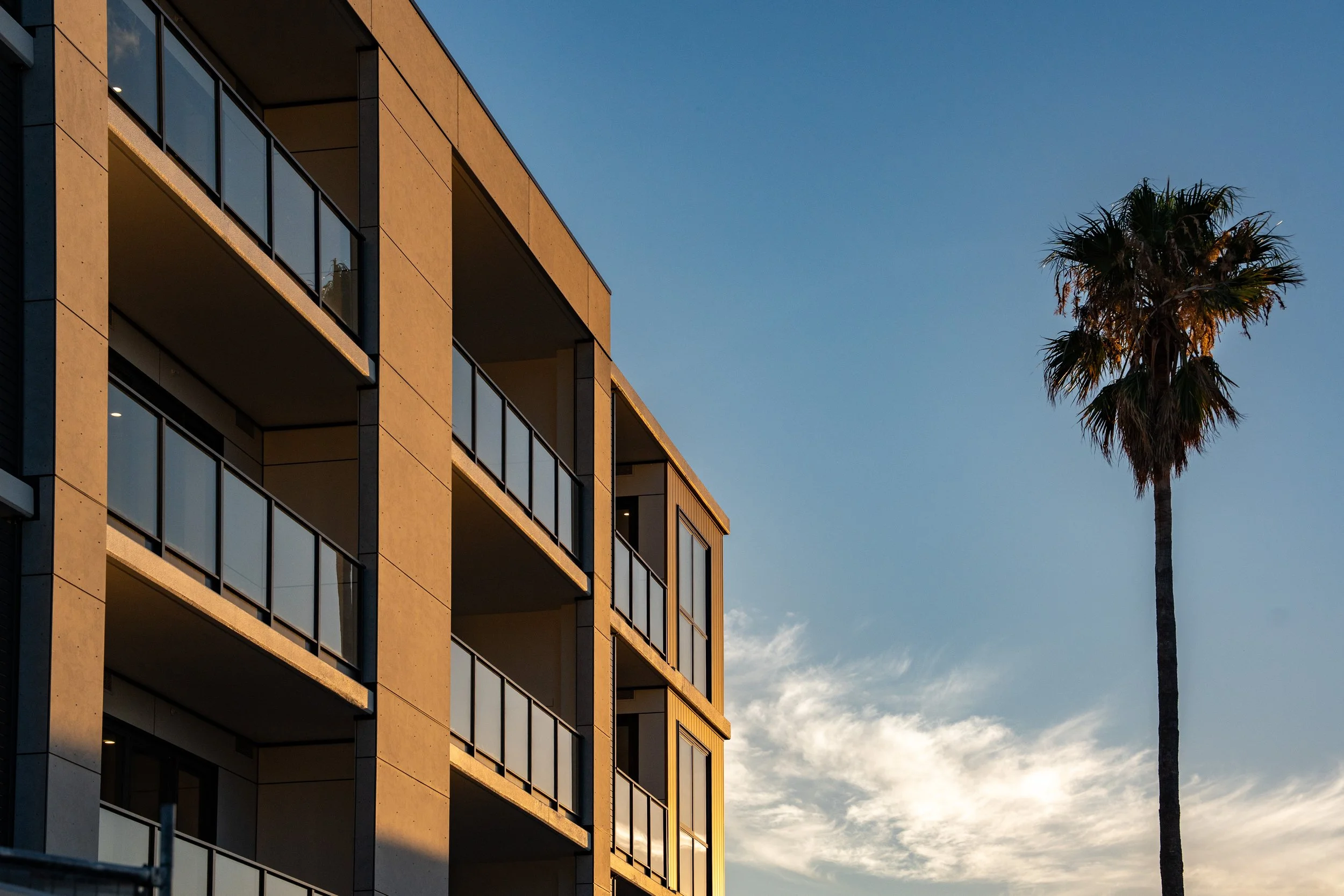 A multi-story modern apartment building with glass balconies and beige exterior walls, next to a tall palm tree under a partly cloudy sky at sunset.