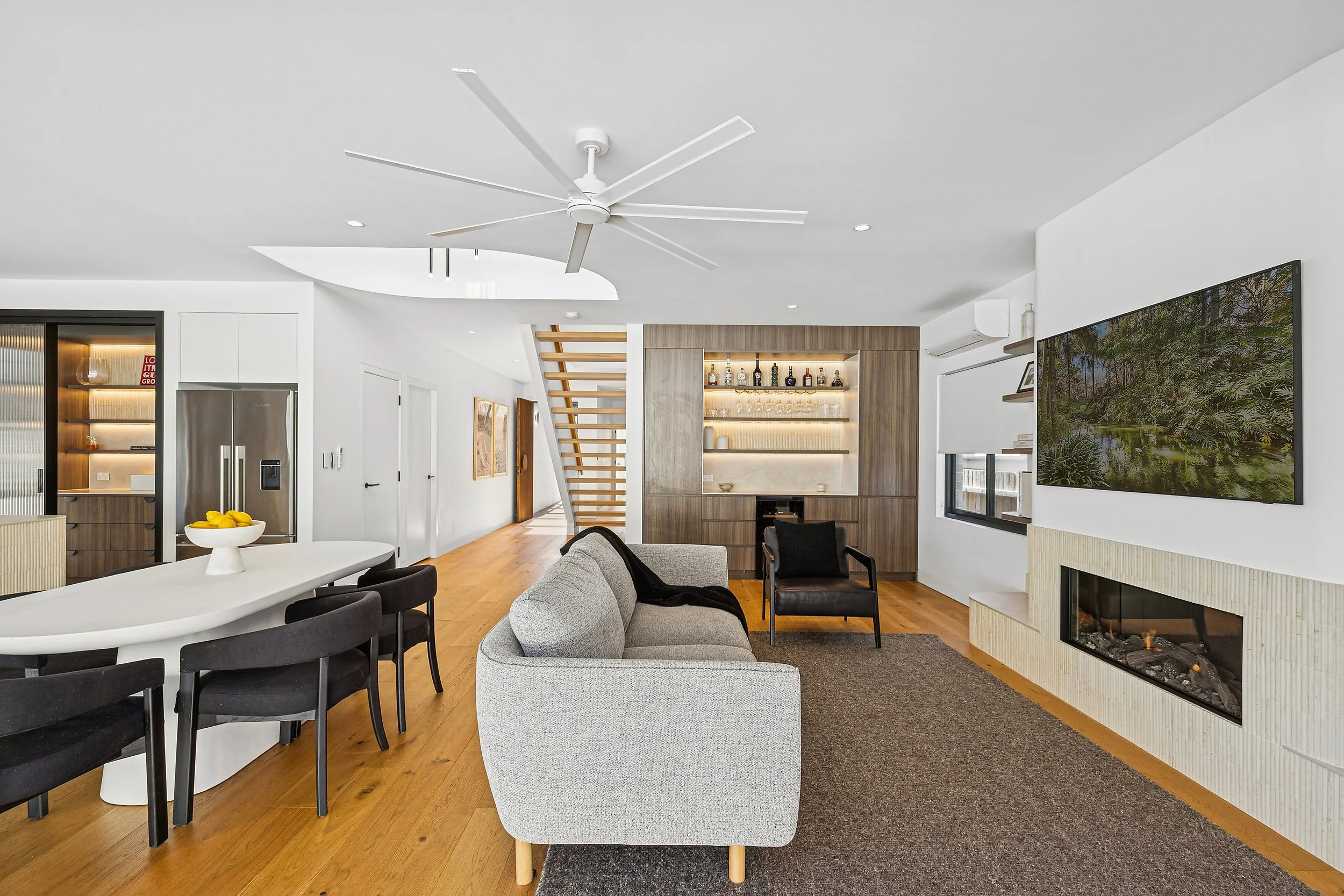 Modern living room with gray couch, black armchair, mounted TV, fireplace, bar area, staircase, and wooden floors