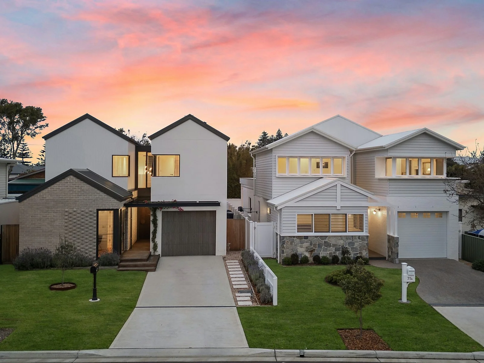 Two modern houses with white exteriors and large windows at sunset, with a green front lawn, trees, and a pink sky in the background.
