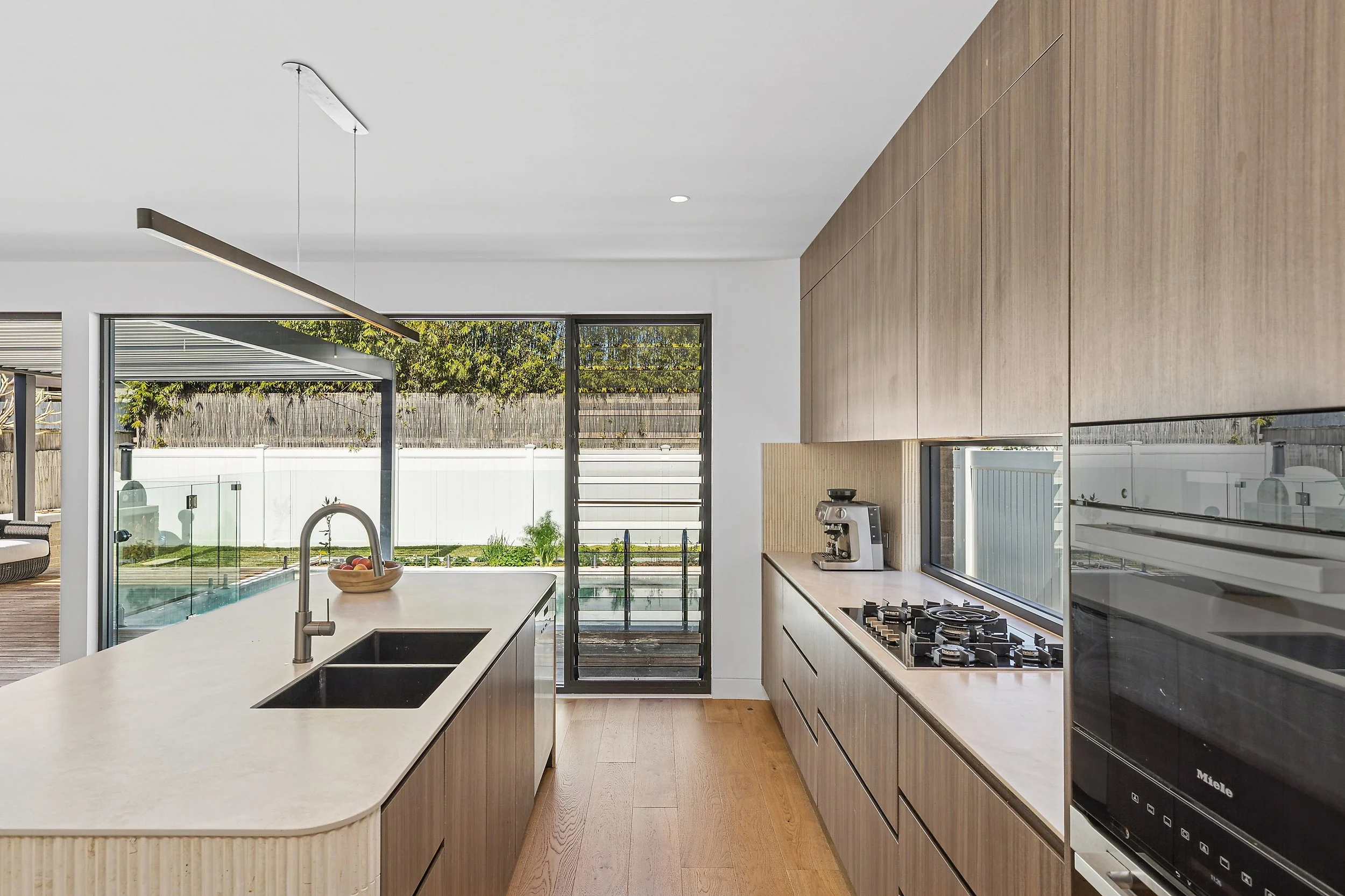 Modern kitchen with light wood cabinets, a white stone countertop, black double sink, coffee maker, gas stove, and built-in oven. Large sliding glass door opens to outdoor patio and backyard with a fence and greenery.
