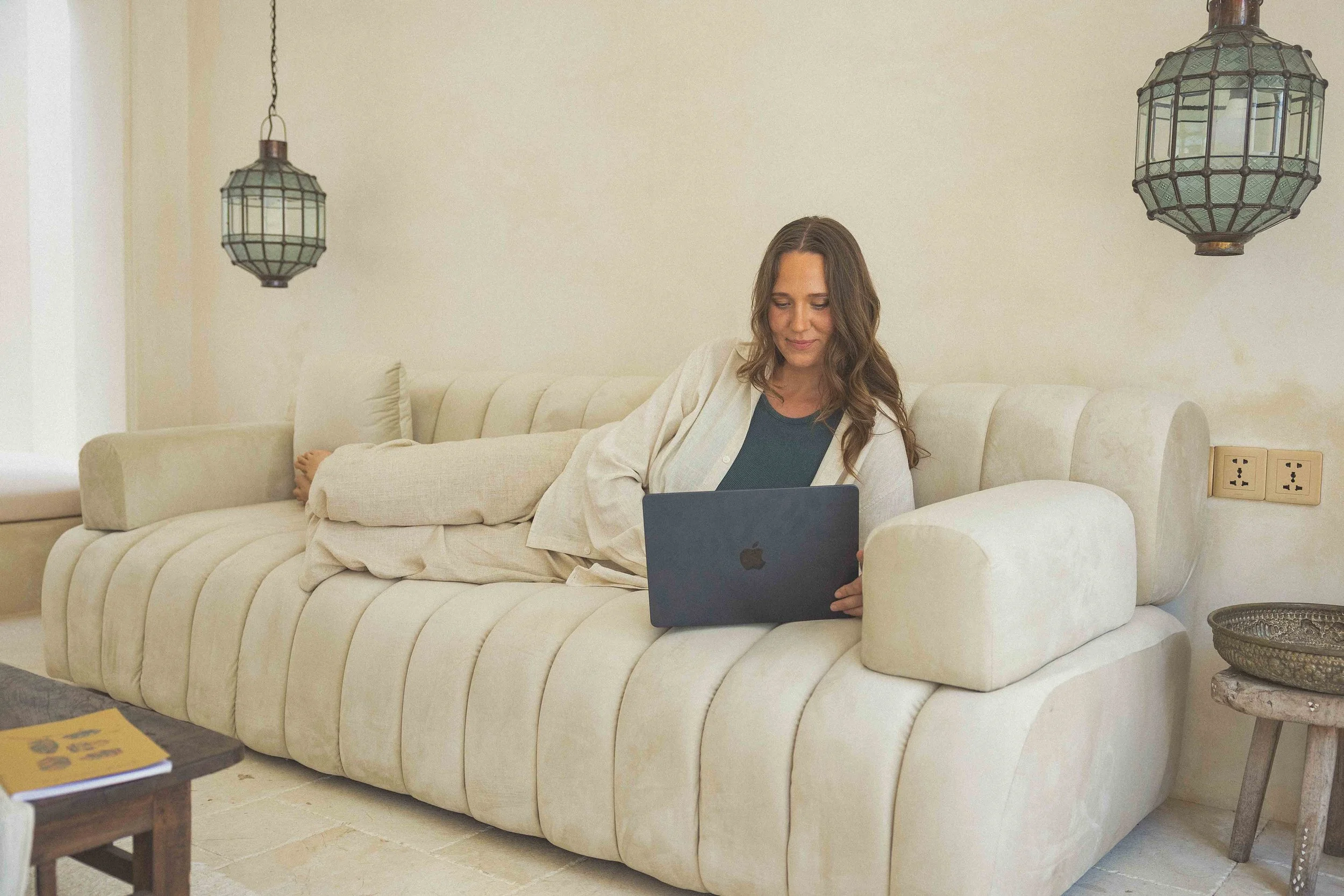 Woman with long wavy hair sitting on a cream-colored sofa, using a laptop, in a cozy living room with hanging lantern lights.