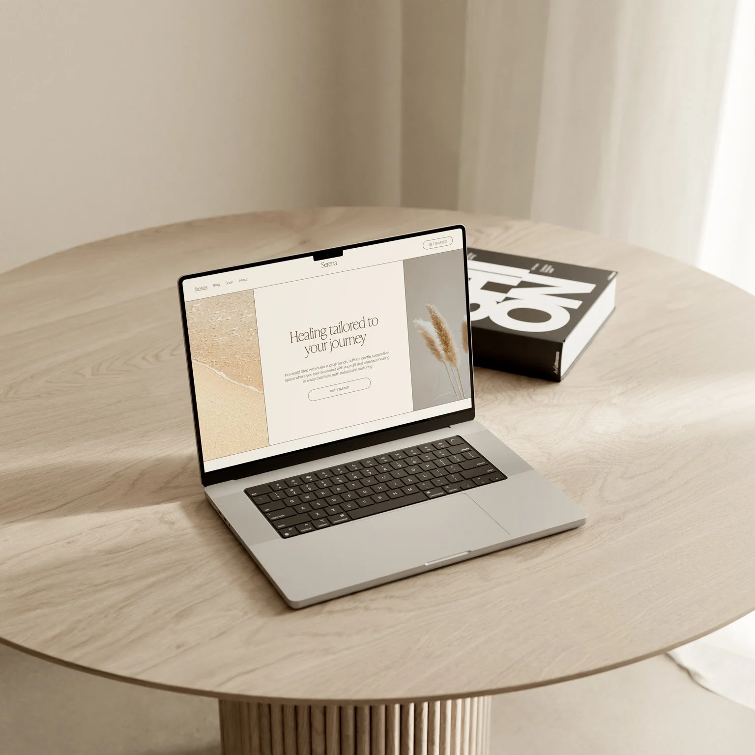 A laptop on a light wood round table displaying a webpage with the headline "Healing tailored to your journey". A book with bold black and white text is placed behind the laptop, and a curtain is partially visible in the background.