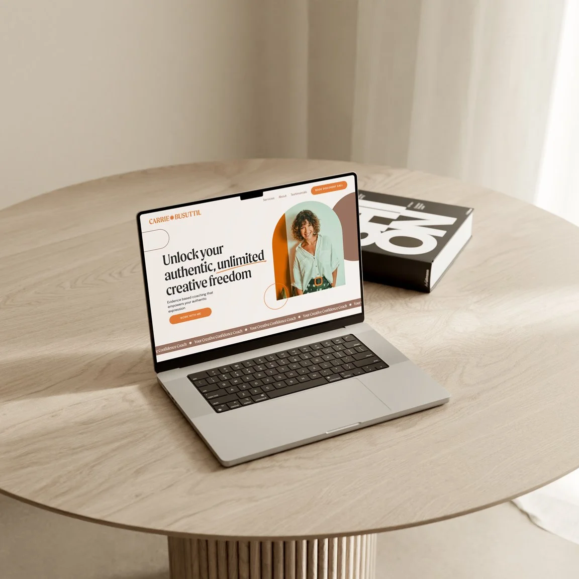Open laptop on a light wood round table, displaying a website for a coaching service with a woman's photo. A closed book is placed behind the laptop.