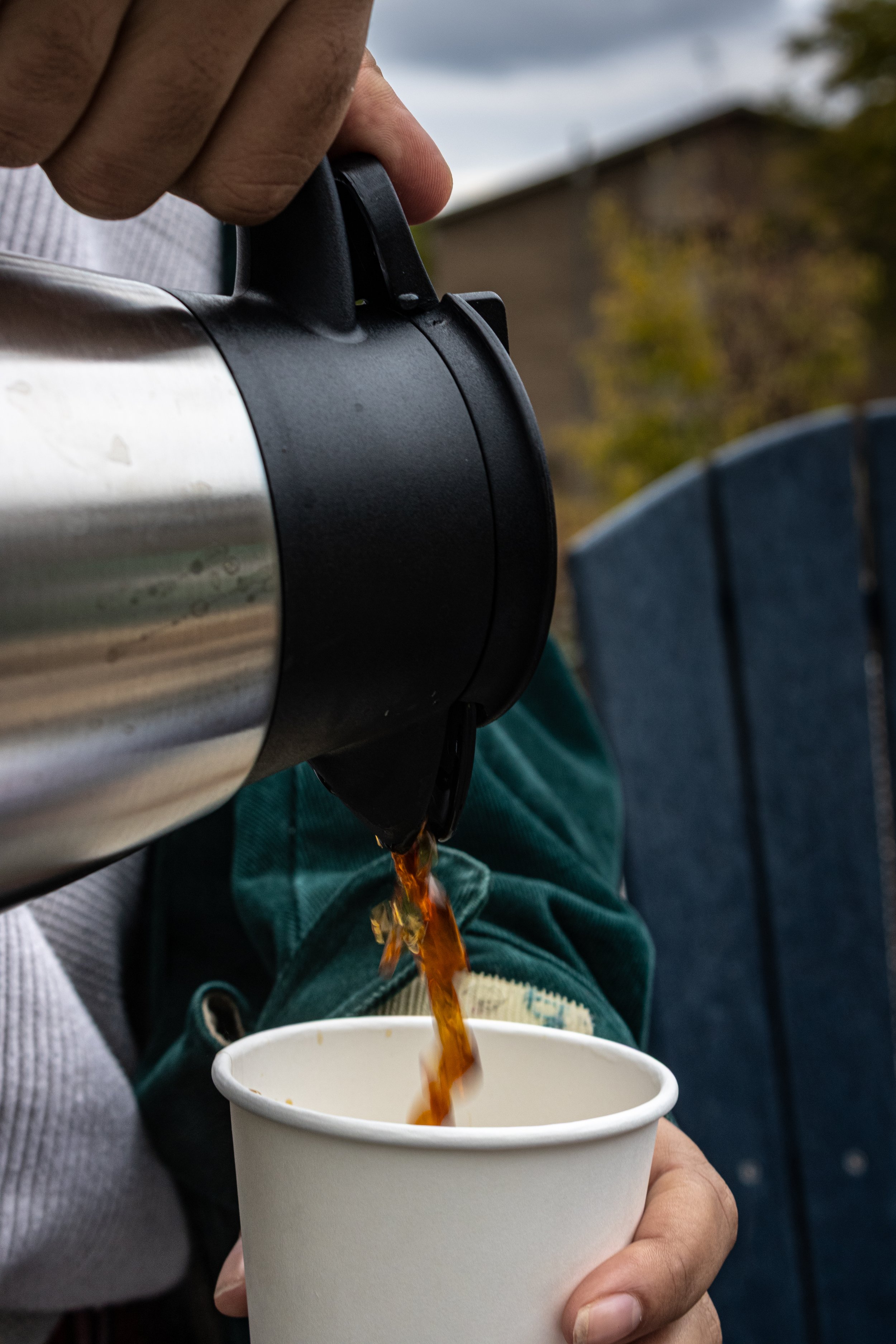 Person pouring hot coffee from a stainless steel thermos into a white paper cup outdoors.
