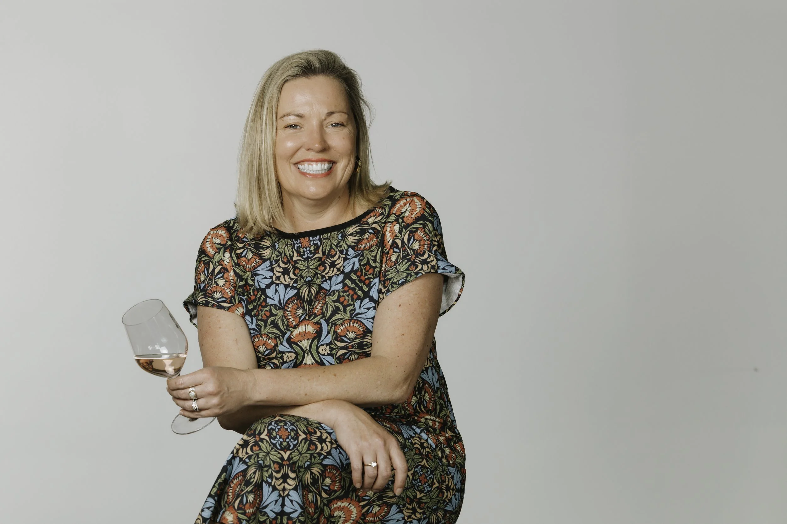 A woman with blonde hair smiling and holding a wine glass of rosé, sitting in front of a plain light gray background.