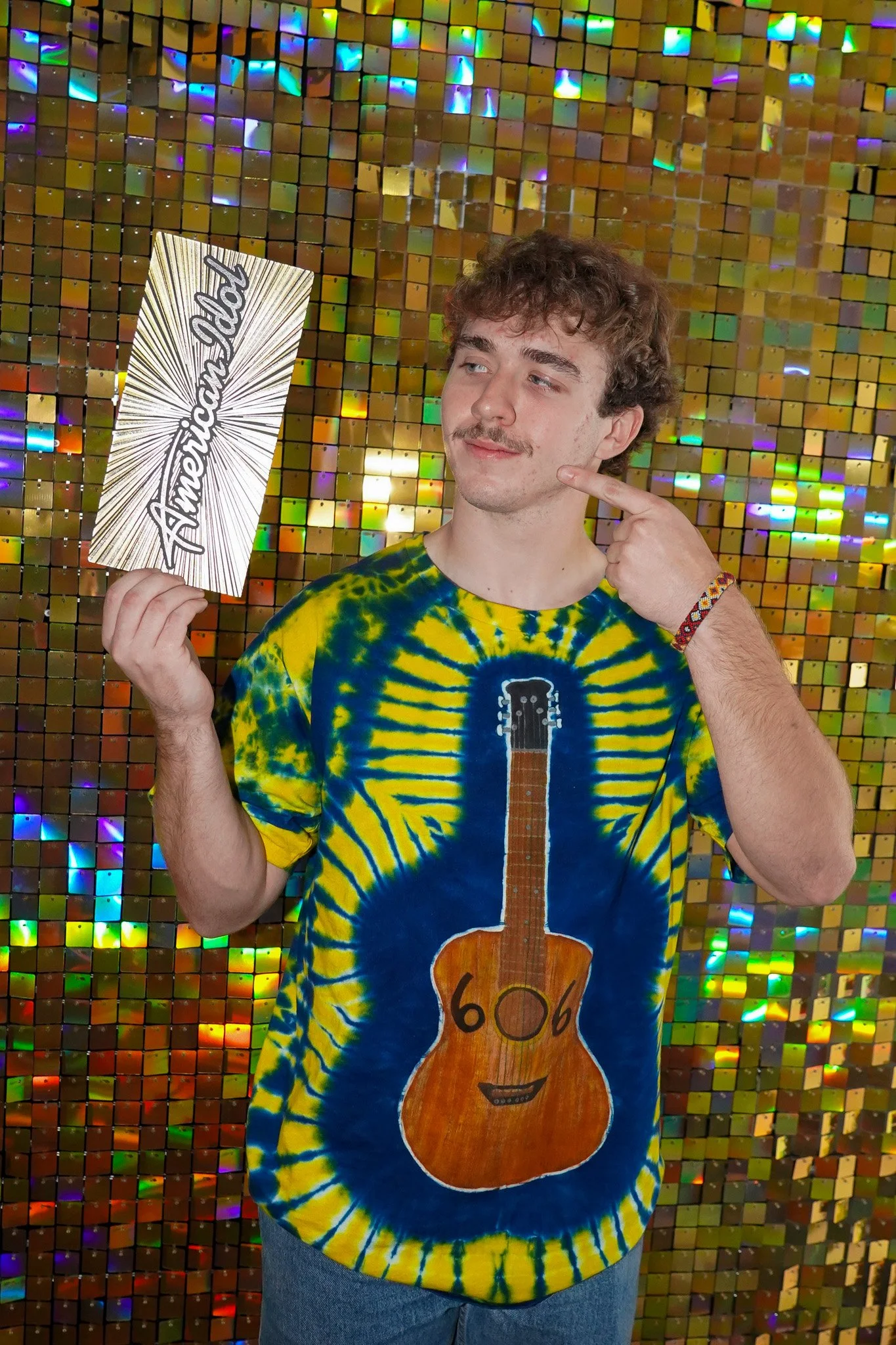 A young man with curly hair and a mustache wearing a colorful tie-dye T-shirt with a guitar illustration and a smiling face. He is holding a sign that says 'American Dad' and pointing to his face, standing in front of a sparkly, multicolored reflective background.