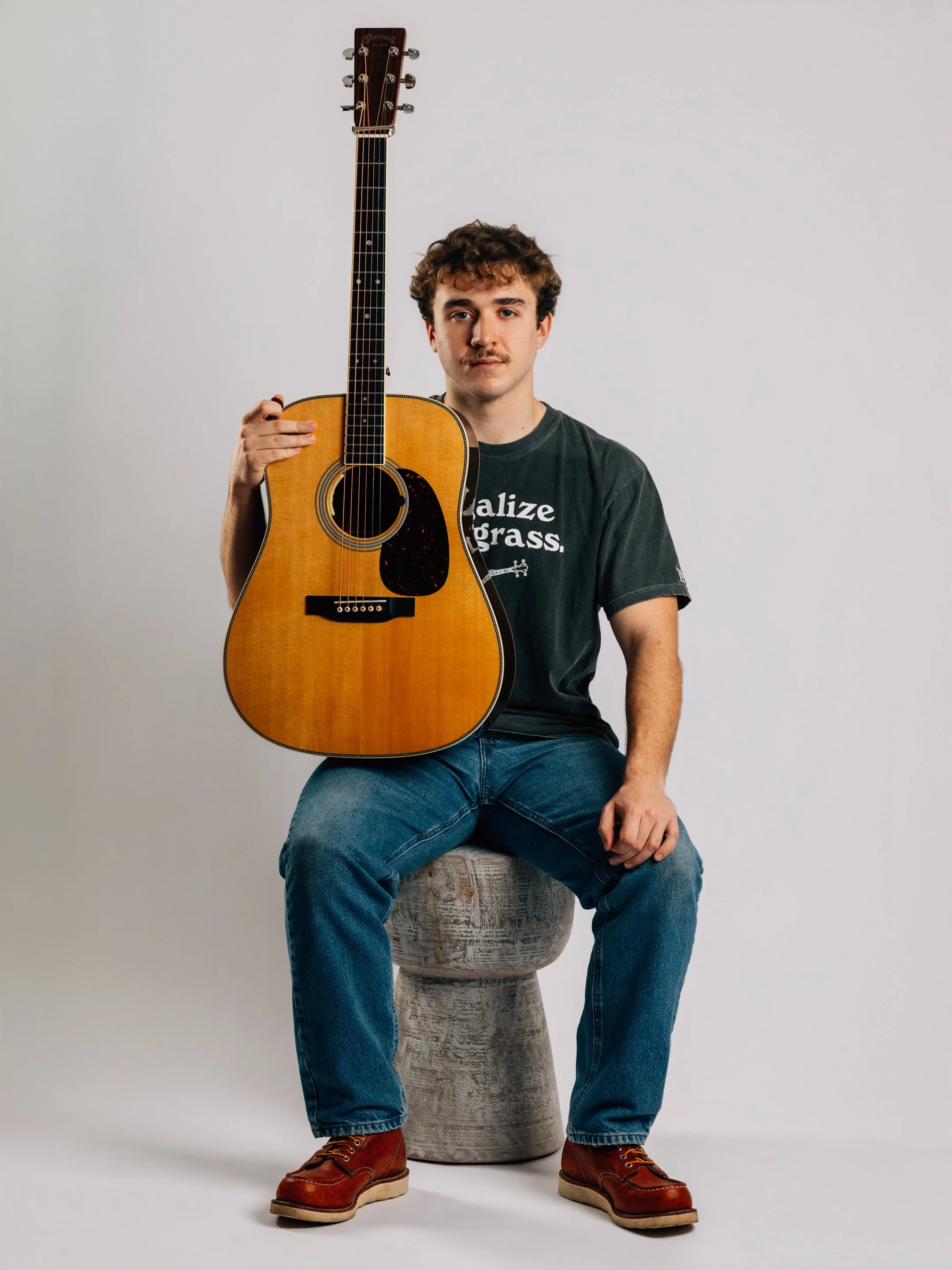 A young man with curly hair and a mustache sitting on a gray stool, holding an acoustic guitar with a natural wood finish and black pickguard, against a plain white background.