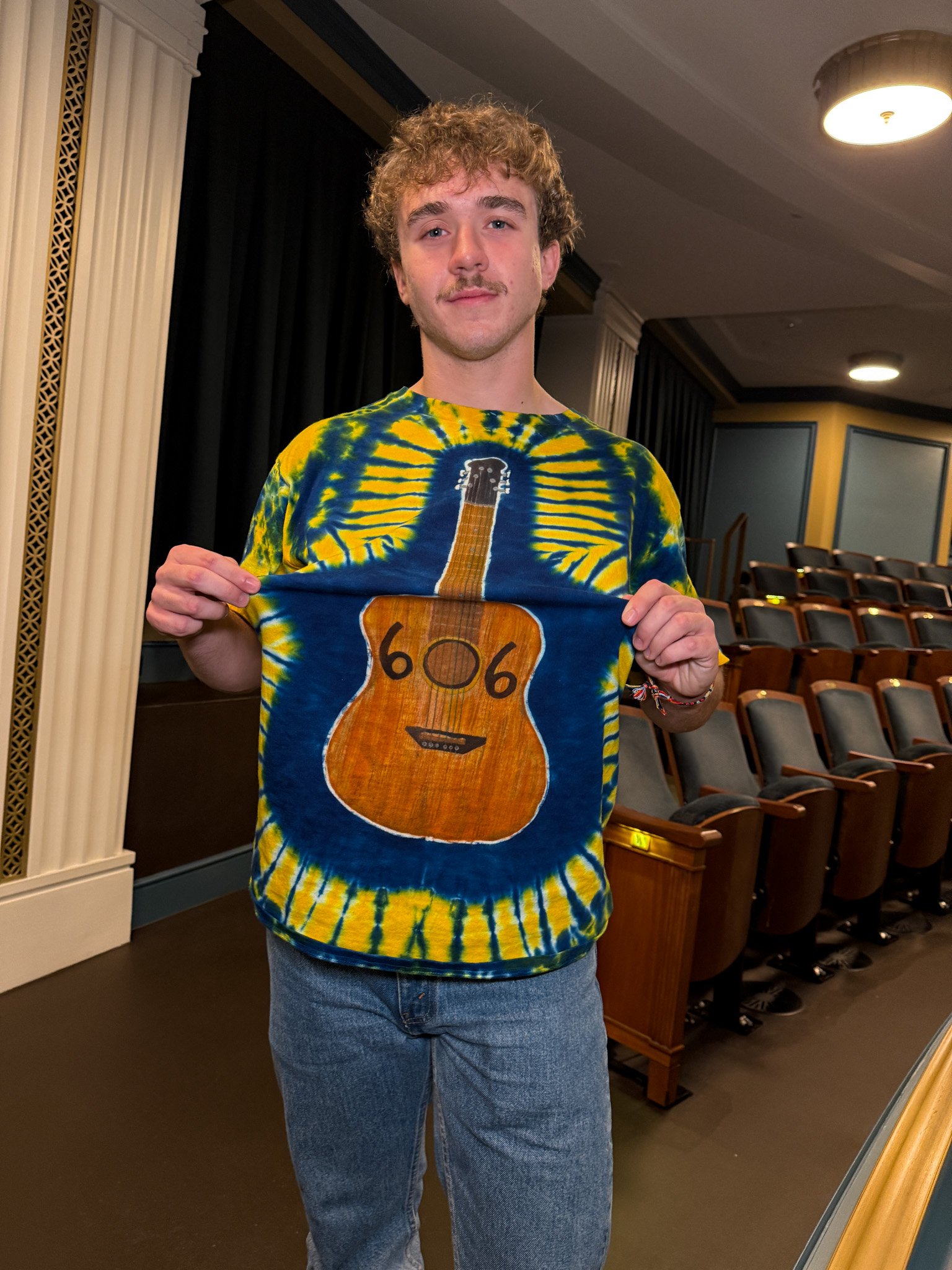 A young man with curly hair and a mustache holding up a colorful t-shirt with a large guitar design and the number 666 on it, standing in an auditorium with empty seats.