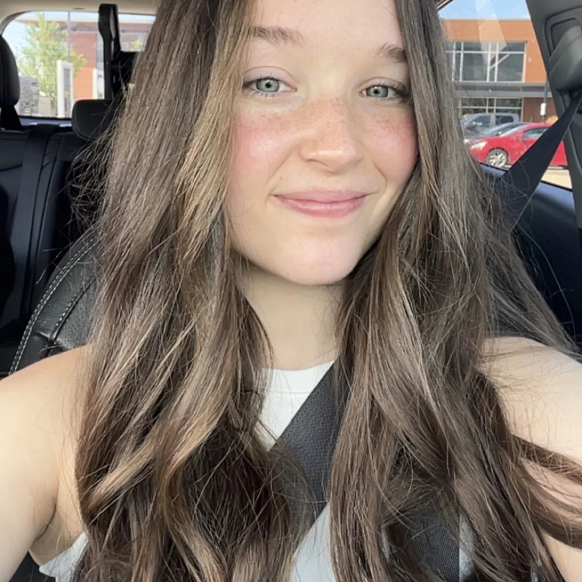 A young woman with long, wavy brown hair and blue eyes taking a selfie inside a car, with a parking lot and red brick building visible through the windows.