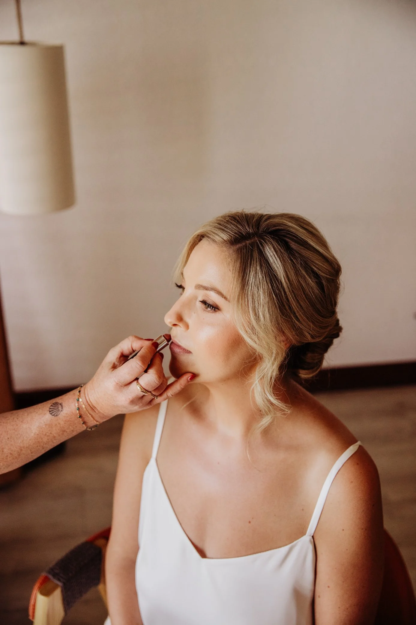 A woman with blonde hair getting her makeup done, sitting in a chair with a white spaghetti strap top on.