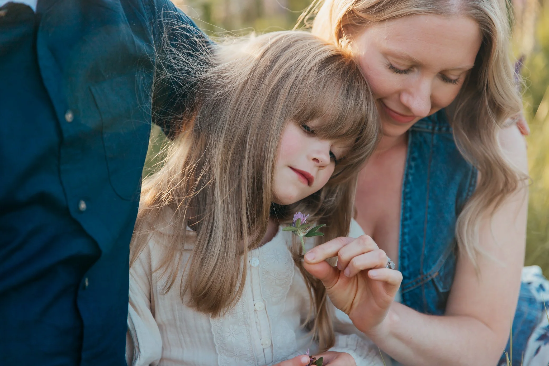 A woman and a girl are closely looking at a small purple flower together outside in nature.